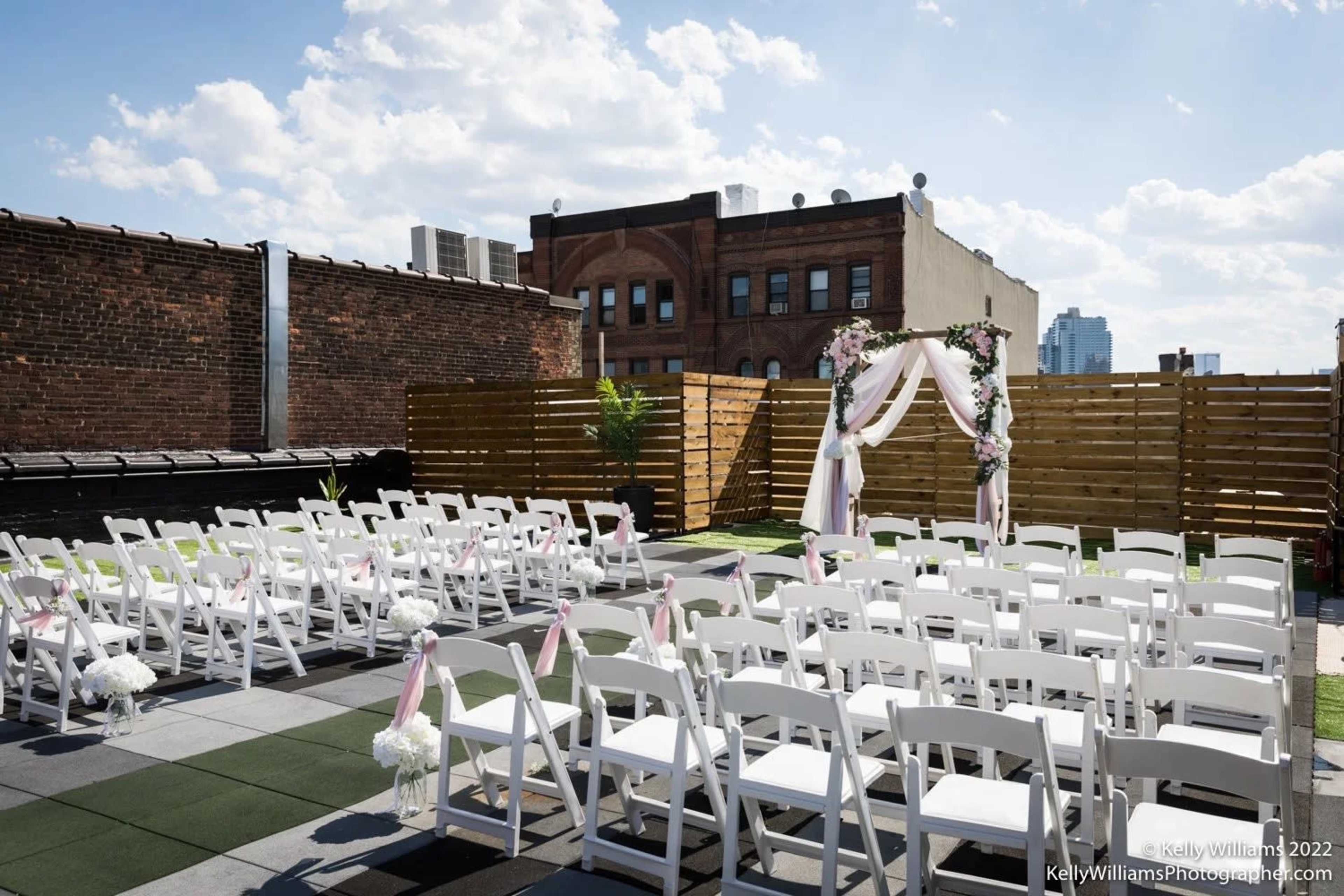 The image shows a rooftop wedding ceremony setup with white chairs arranged in rows facing a floral arch.