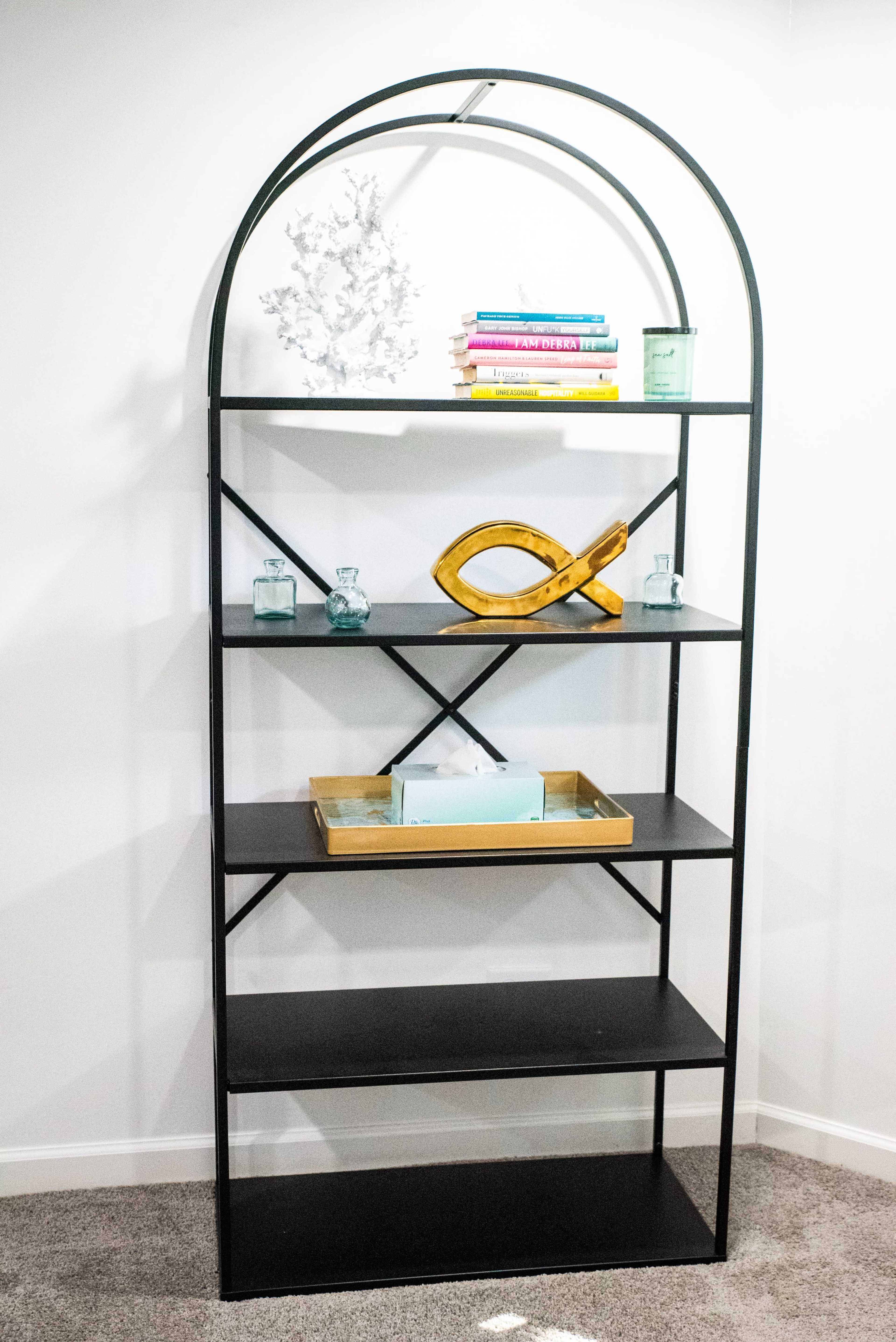 A black metal shelf unit with arch-shaped top, featuring multiple shelves displaying books, decorative items, and glass containers against a white wall.