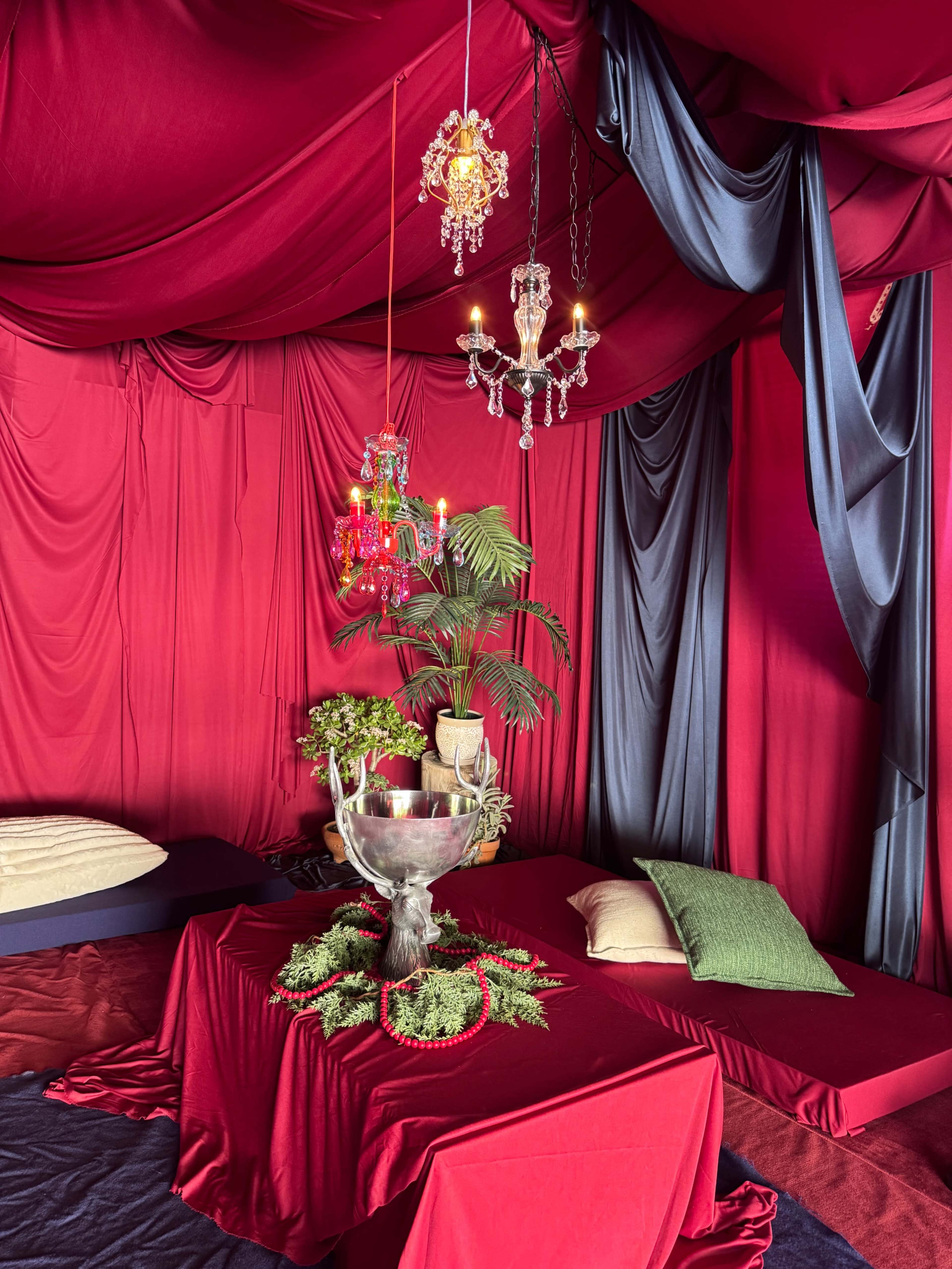 The image shows an intimate setup with a red draped tent featuring a table at its center adorned with greenery and decorative objects, alongside a chandelier hanging from the ceiling.