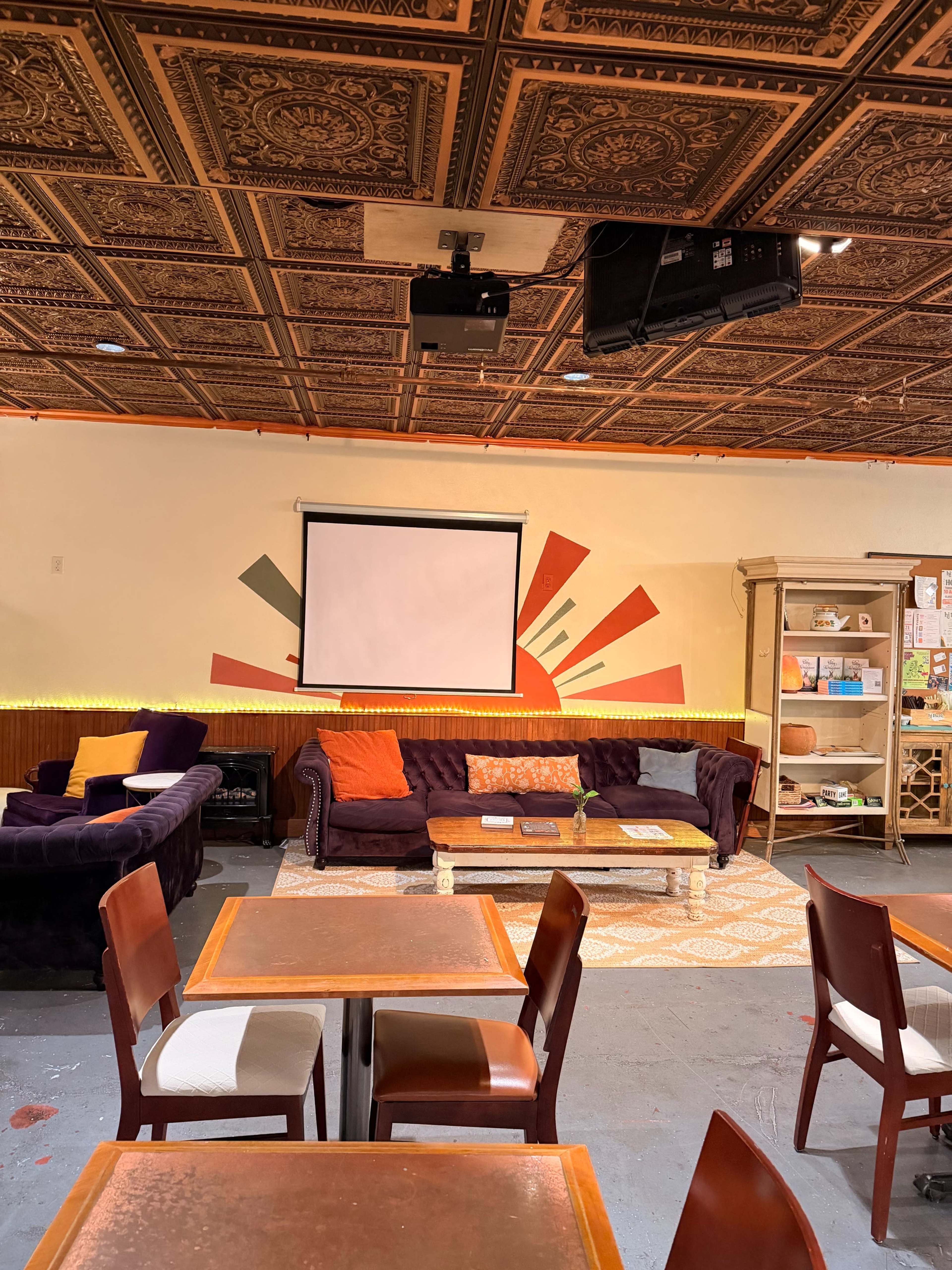 The image shows a cozy café interior with a projector screen on the wall, a seating area featuring purple couches, and wooden tables and chairs.
