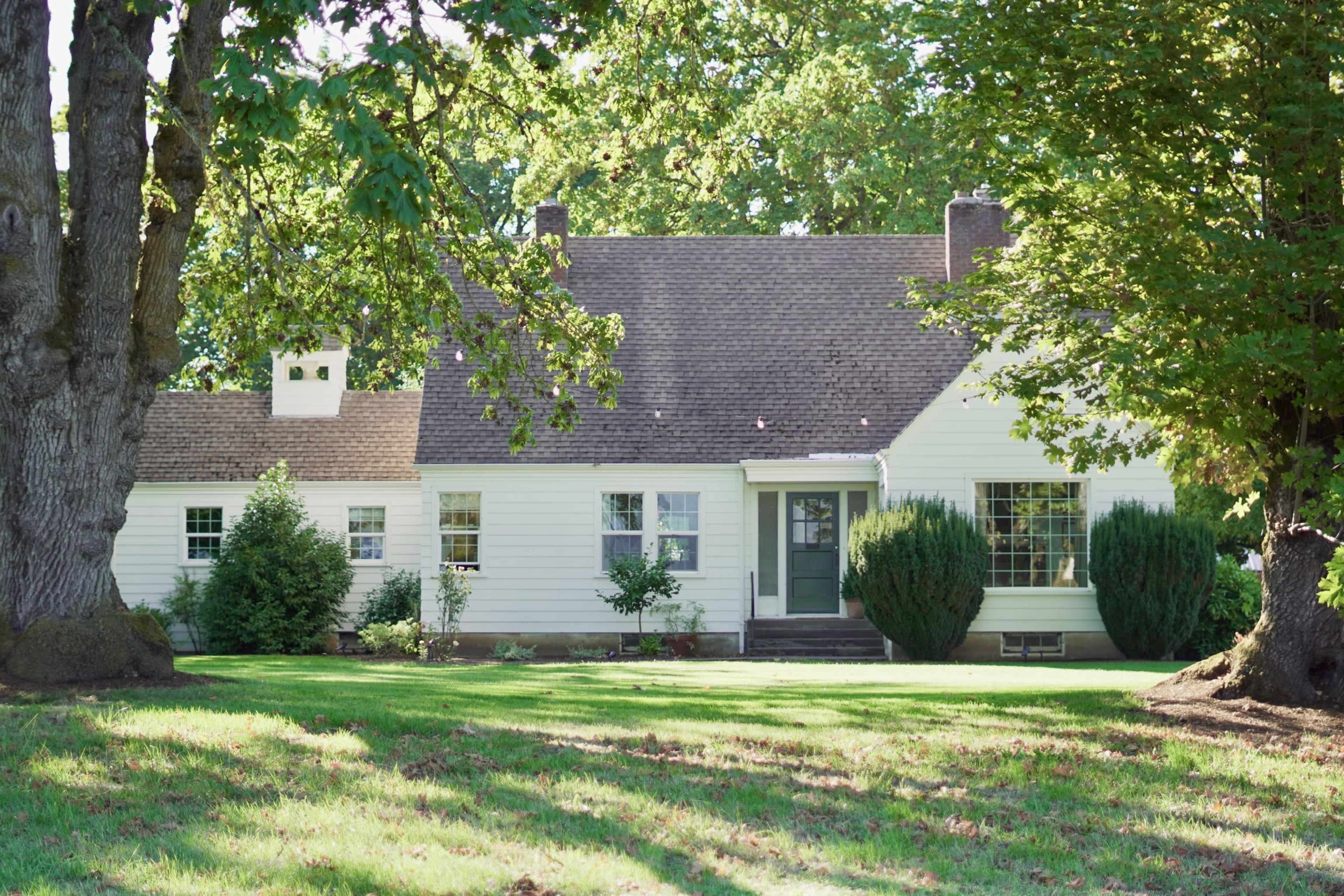 A white, single-story house with a steep roof and green accents is surrounded by tall trees and a lawn.