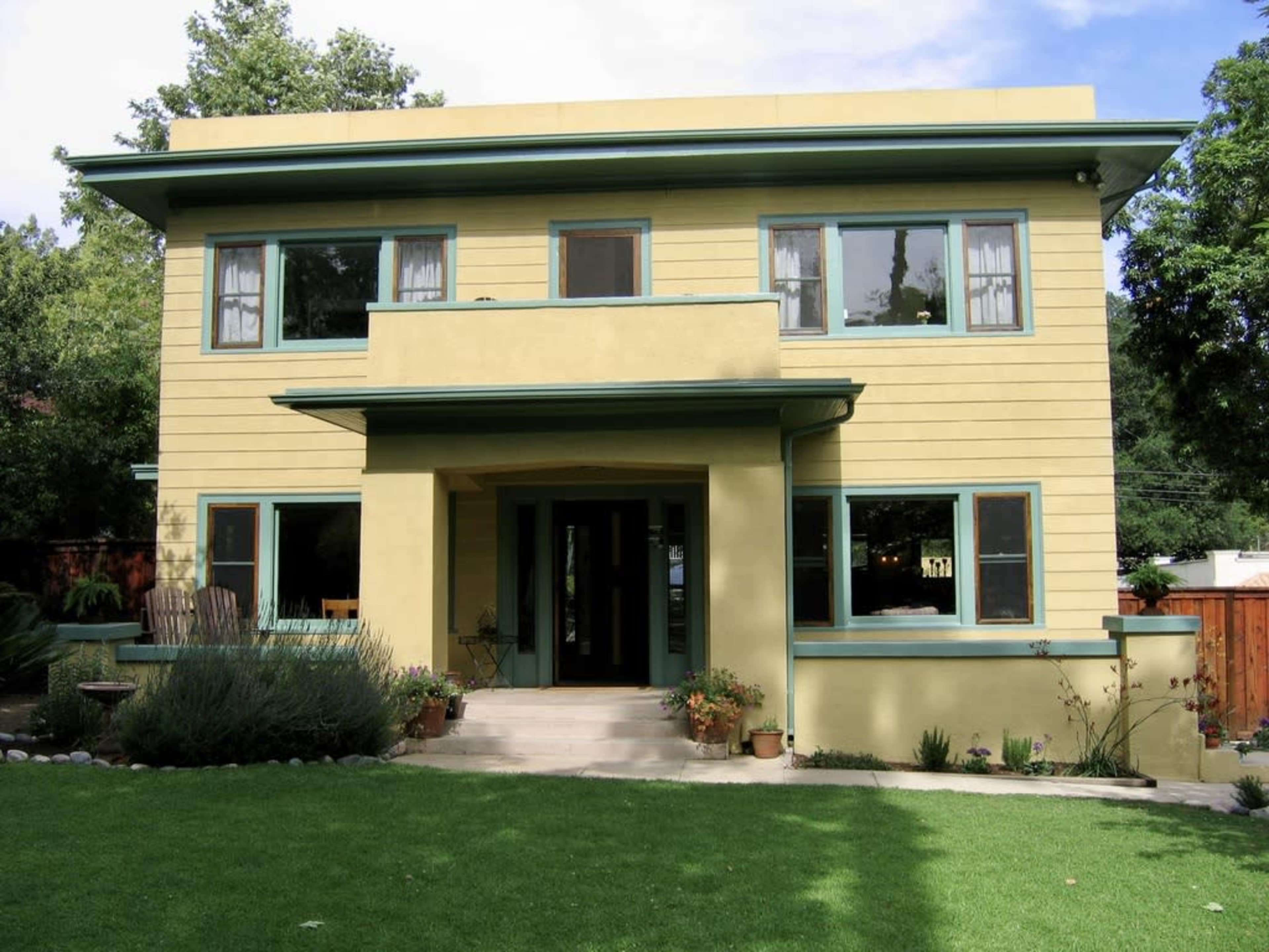 A two-story yellow house with green window frames, a front porch, and a well-maintained lawn.