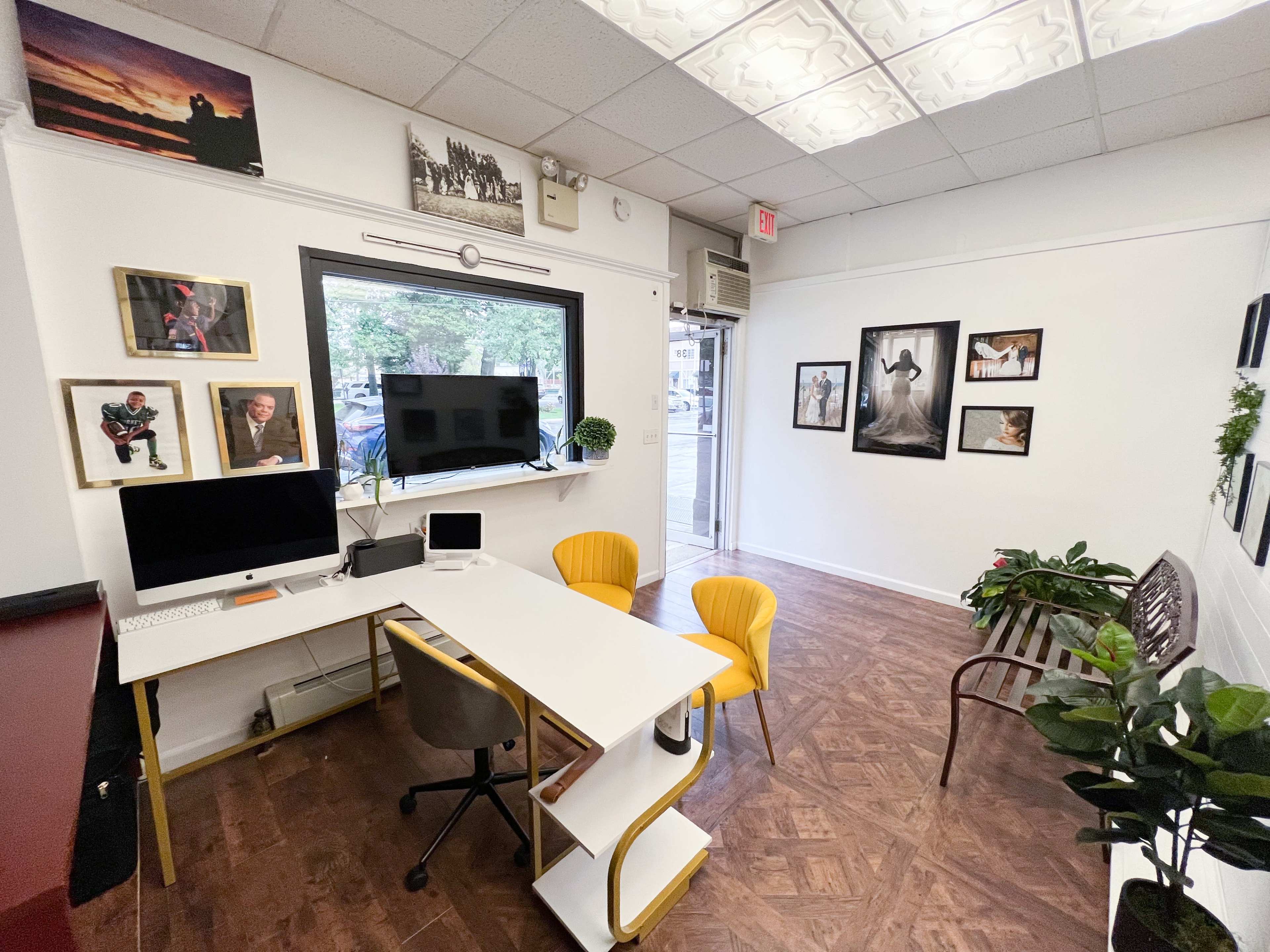 A small office space featuring a white desk, yellow chairs, a computer, framed photographs on the walls, and plants in the corner.