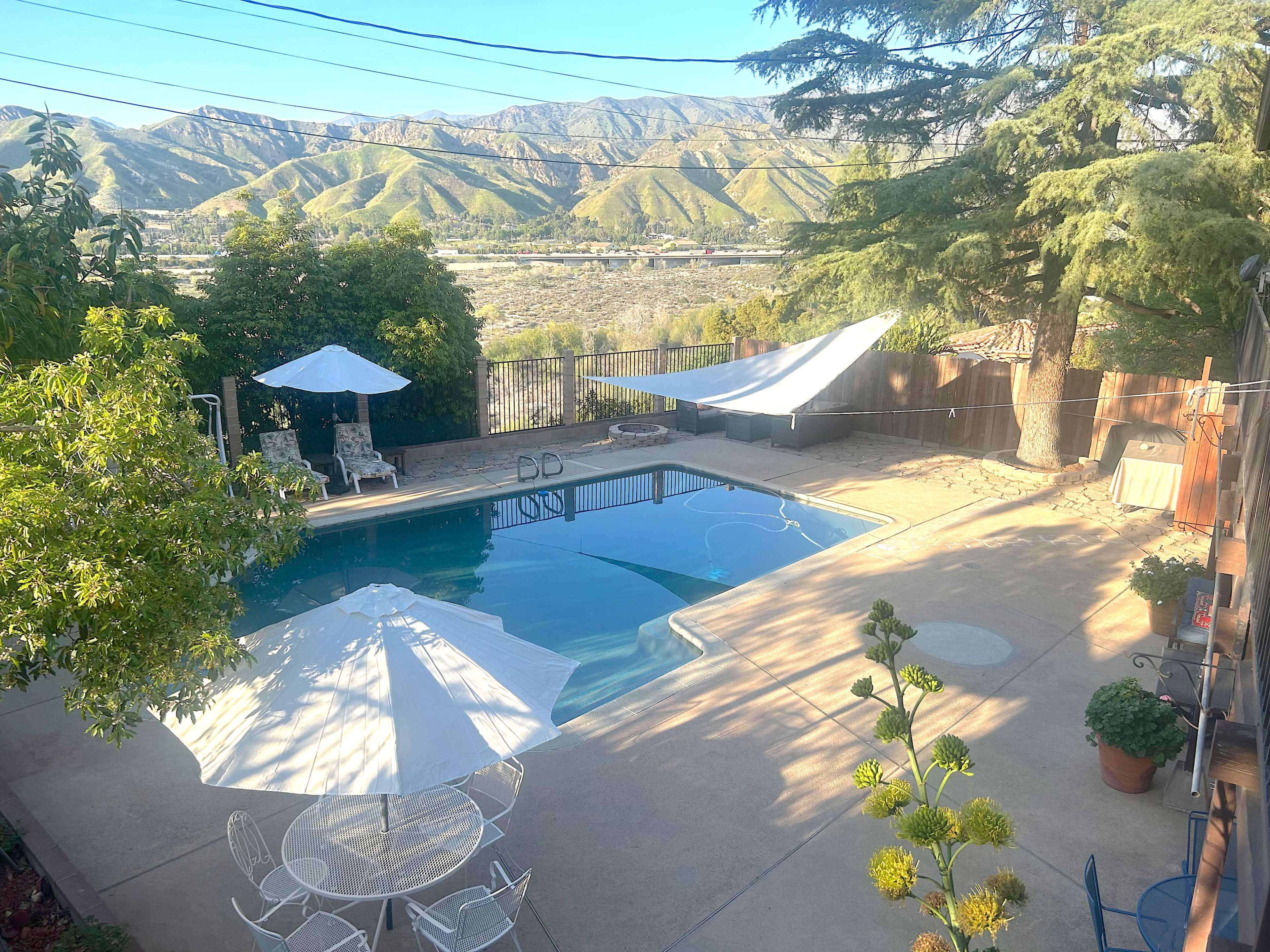 A swimming pool surrounded by a concrete patio, lawn chairs, and umbrellas, with mountains visible in the background.