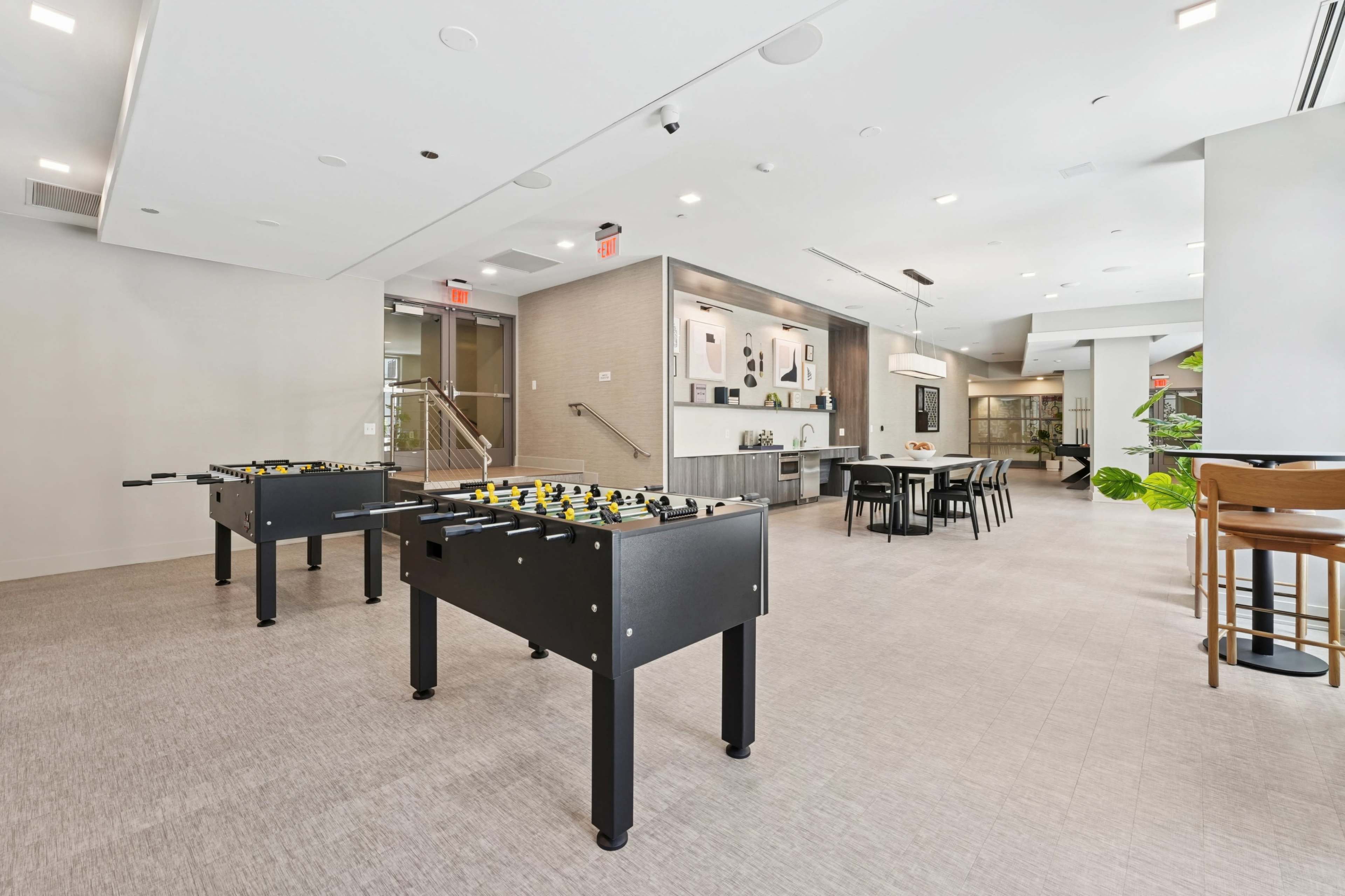 The image shows a modern recreational area featuring two foosball tables and a dining space with a table and chairs.