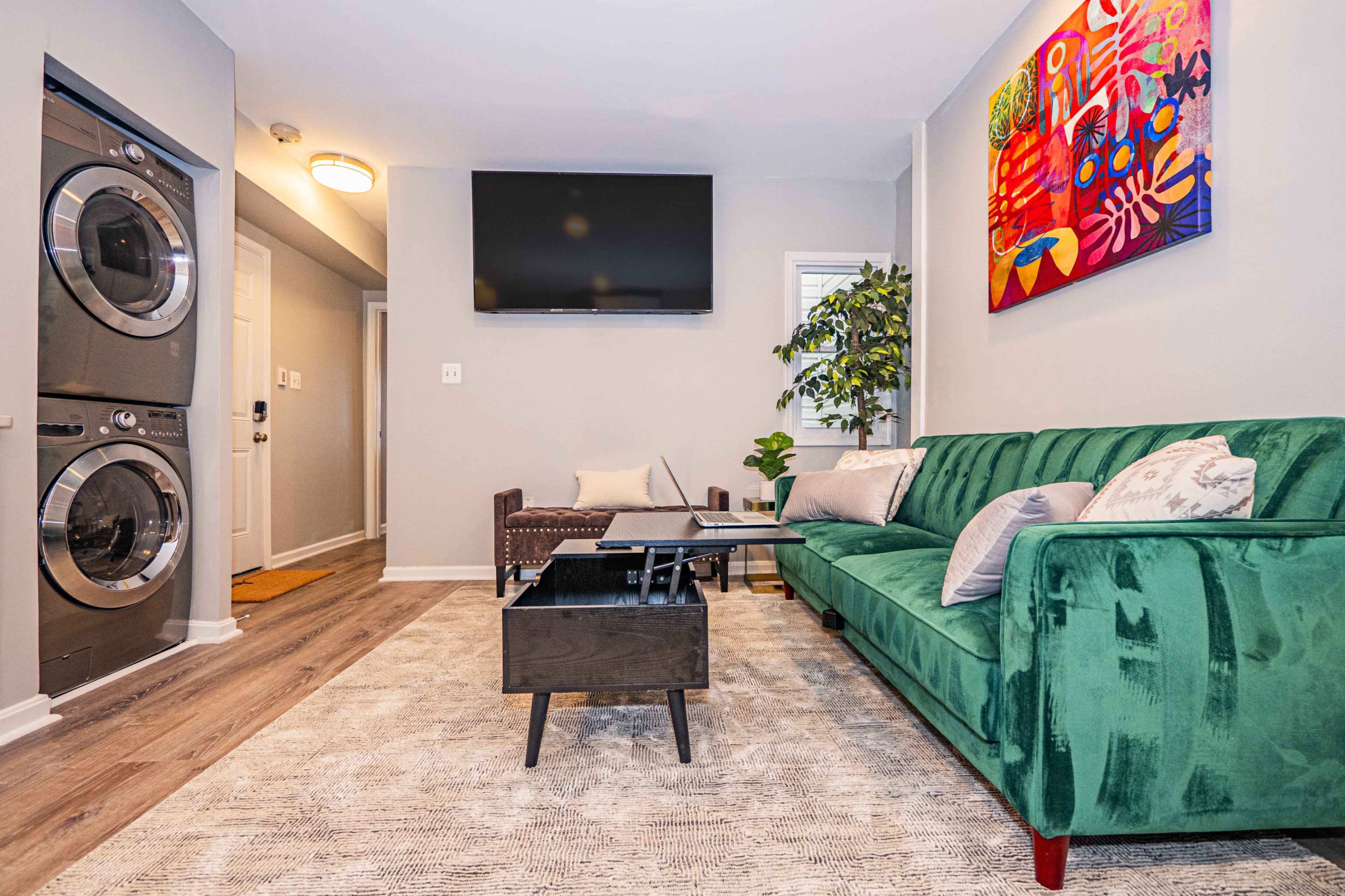 The image shows a modern living room with a green sofa, a coffee table, a wall-mounted TV, and a stackable washer-dryer in the corner.