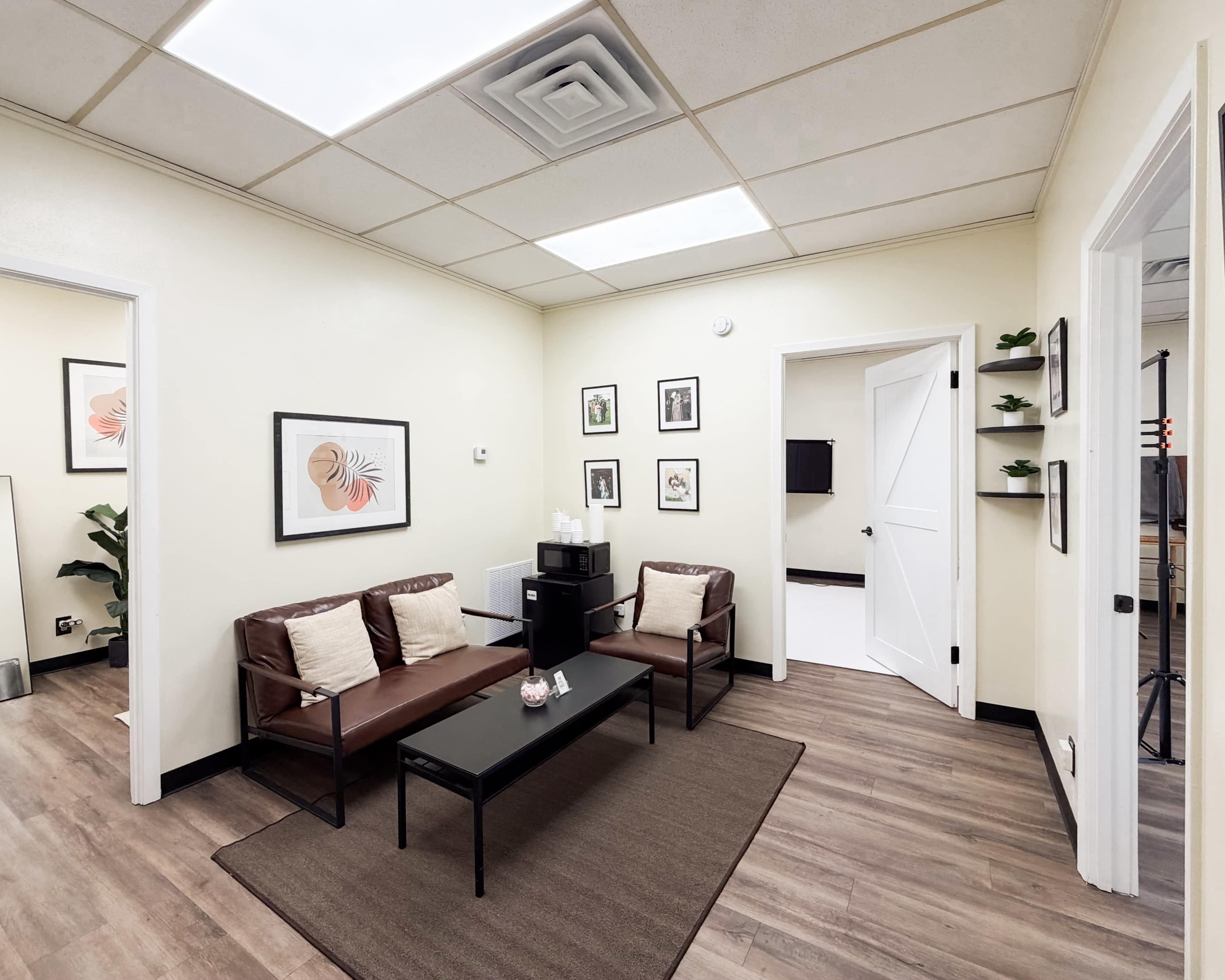 The image shows a small waiting area featuring two brown leather chairs, a black coffee table, framed artwork on the walls, and a plant in the corner.