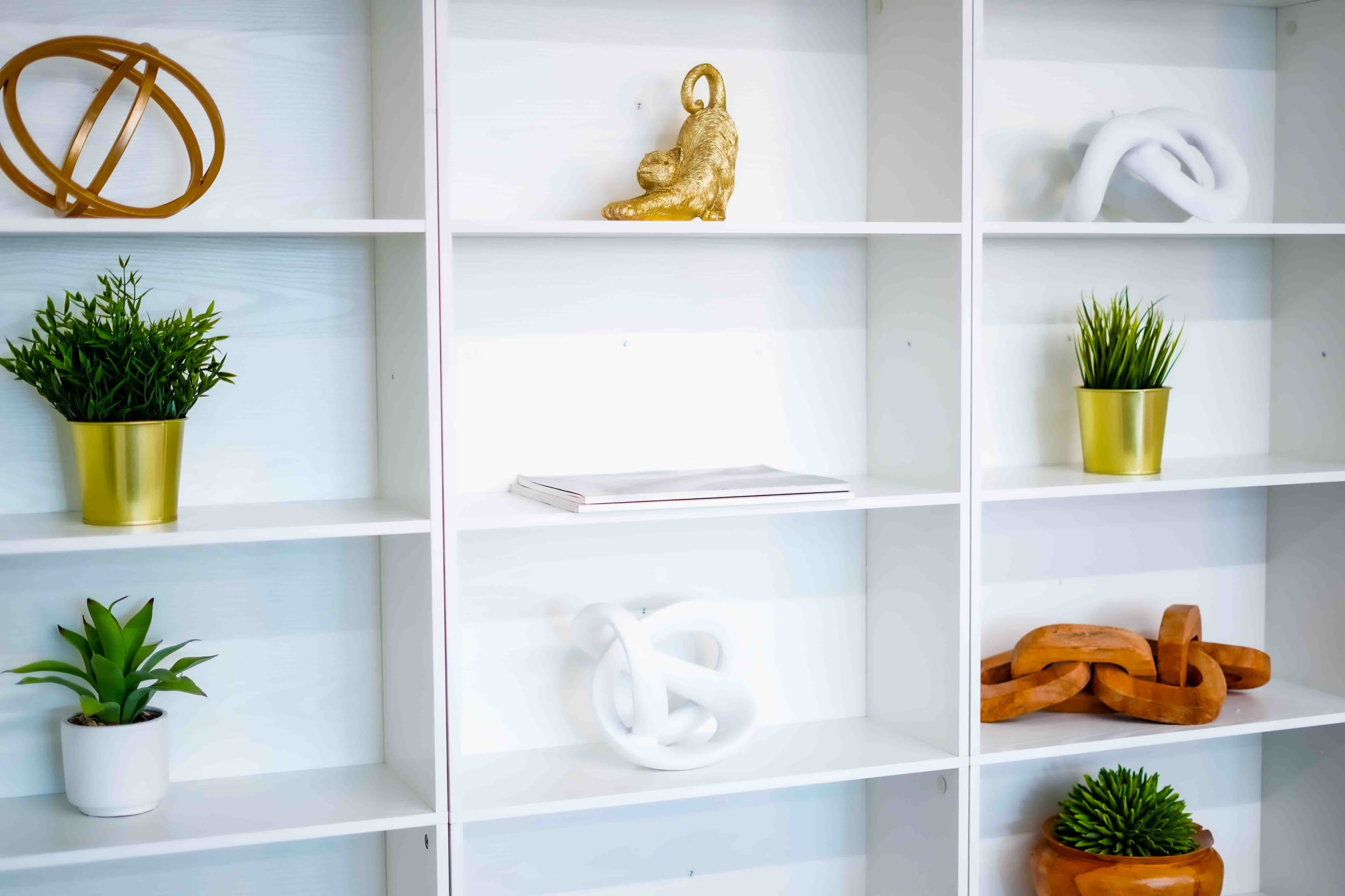The image shows a white shelving unit containing various decorative items, including sculptures, potted plants, and a magazine.