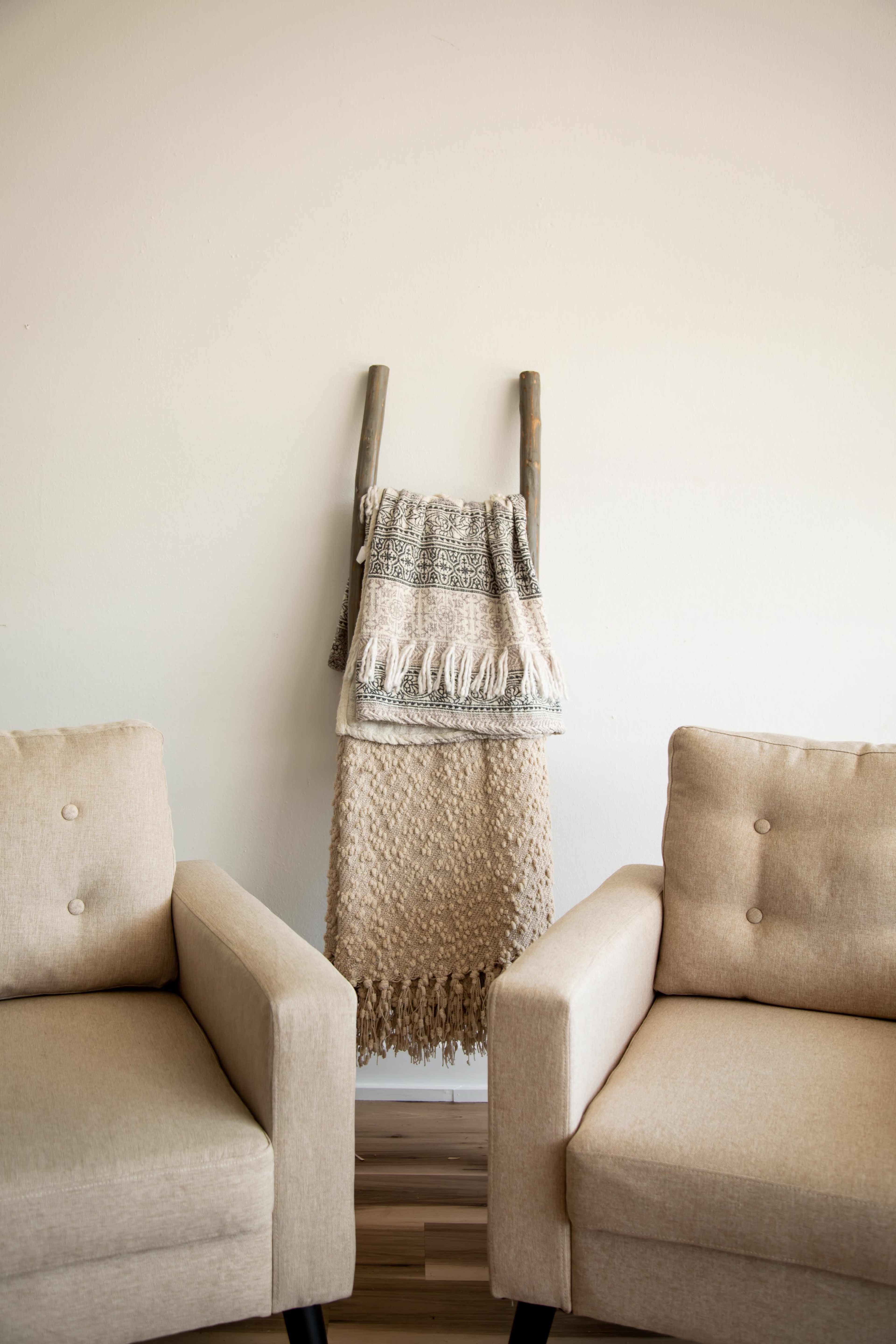 A woven blanket hangs on a wooden ladder between two beige armchairs.