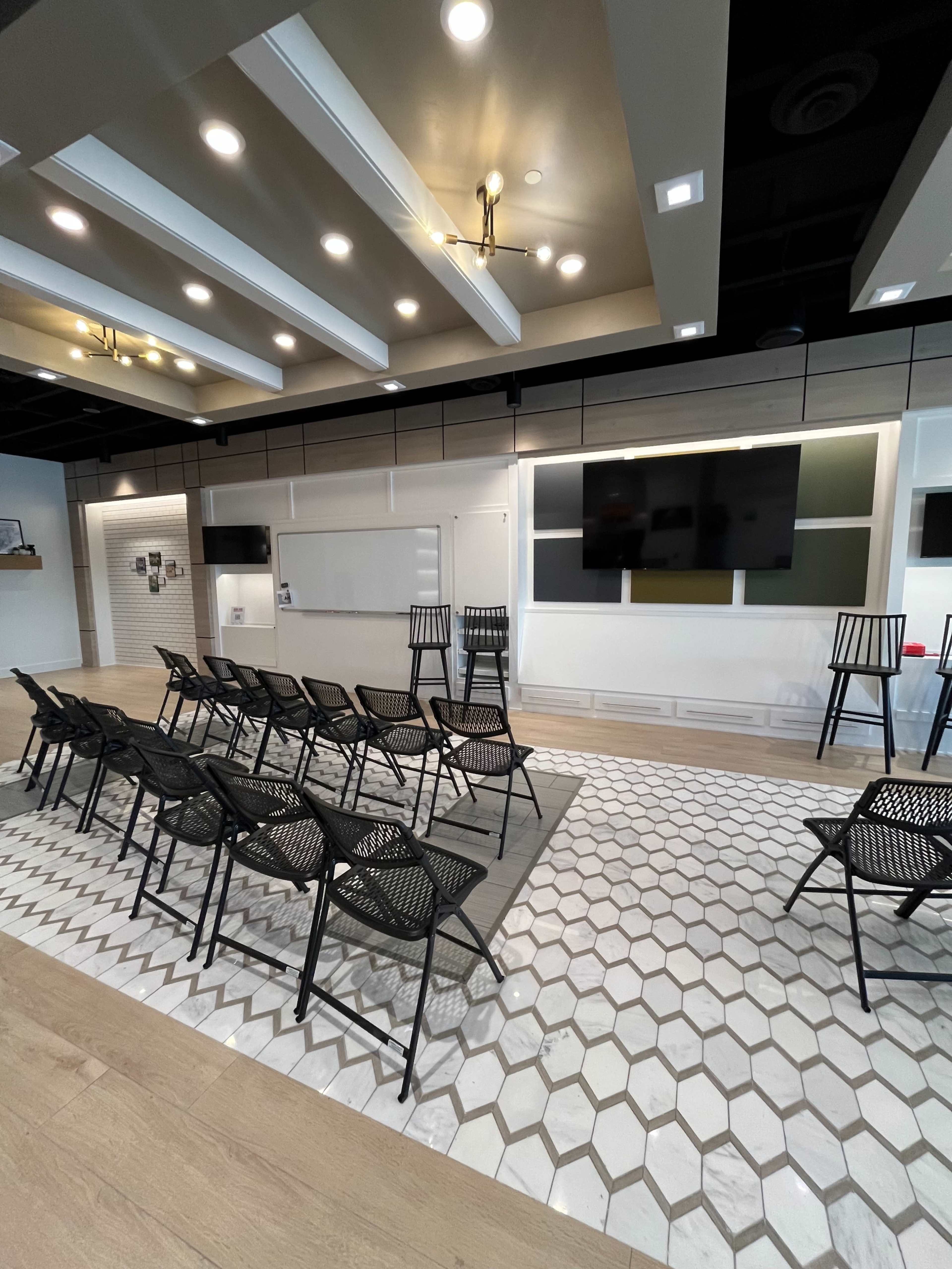 The image shows a modern meeting space with rows of black chairs facing a wall equipped with two large screens and whiteboards, surrounded by patterned tiled flooring.
