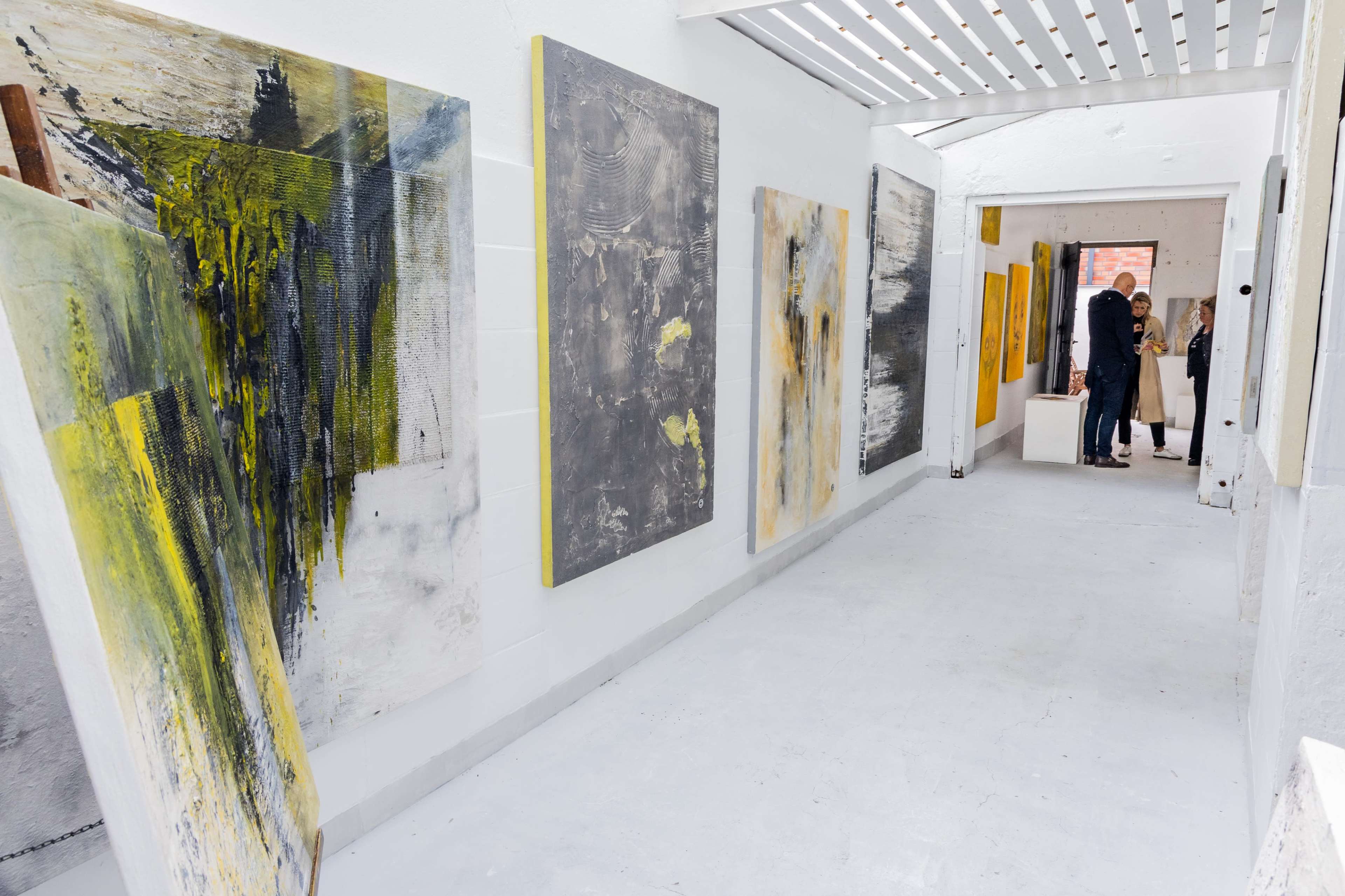 The image depicts an interior gallery space with various large abstract paintings displayed on the white walls and visitors engaging in conversation at the end of the corridor.