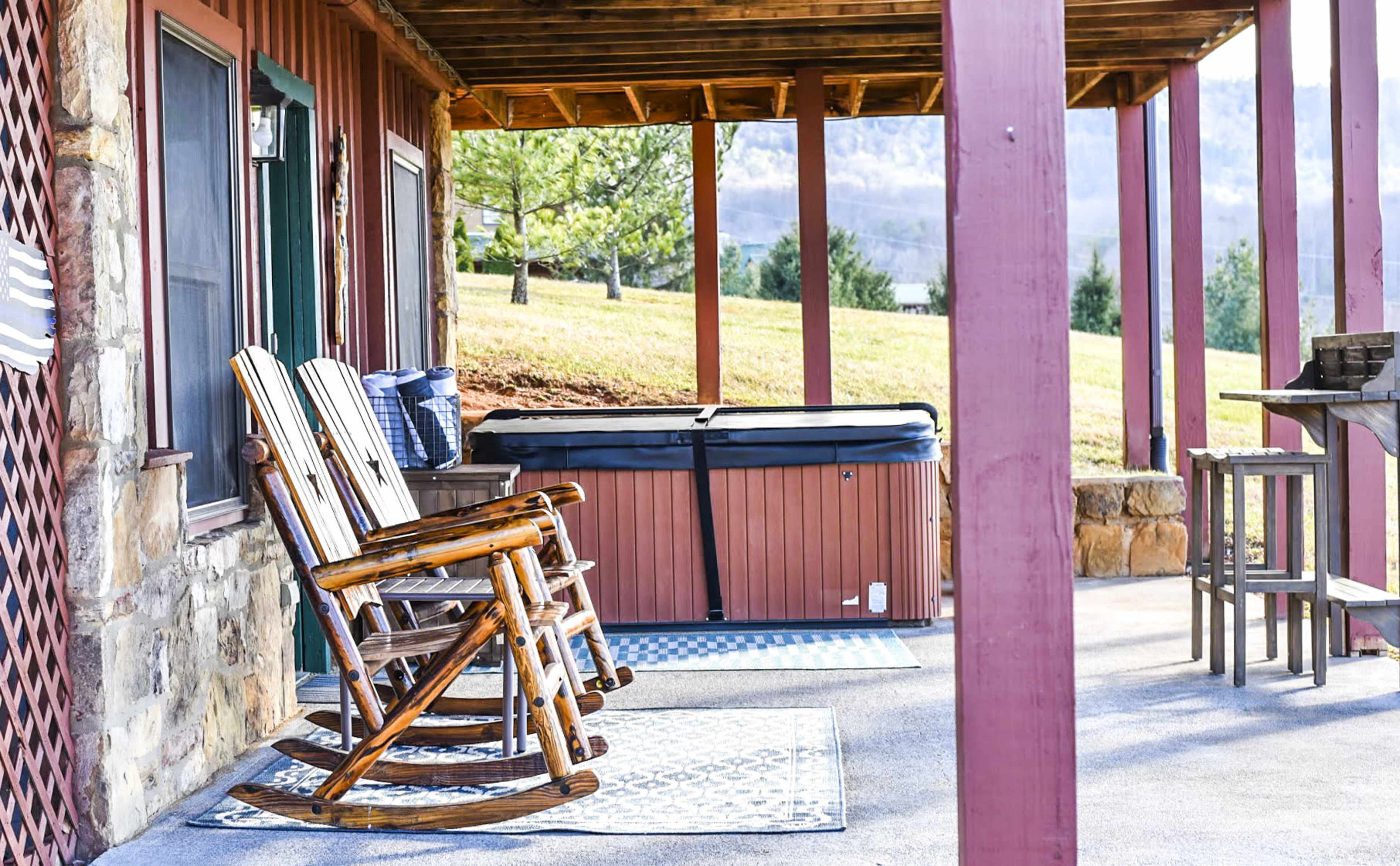The image shows a porch area with two wooden rocking chairs and a hot tub, surrounded by grassy landscaping and mountains in the background.