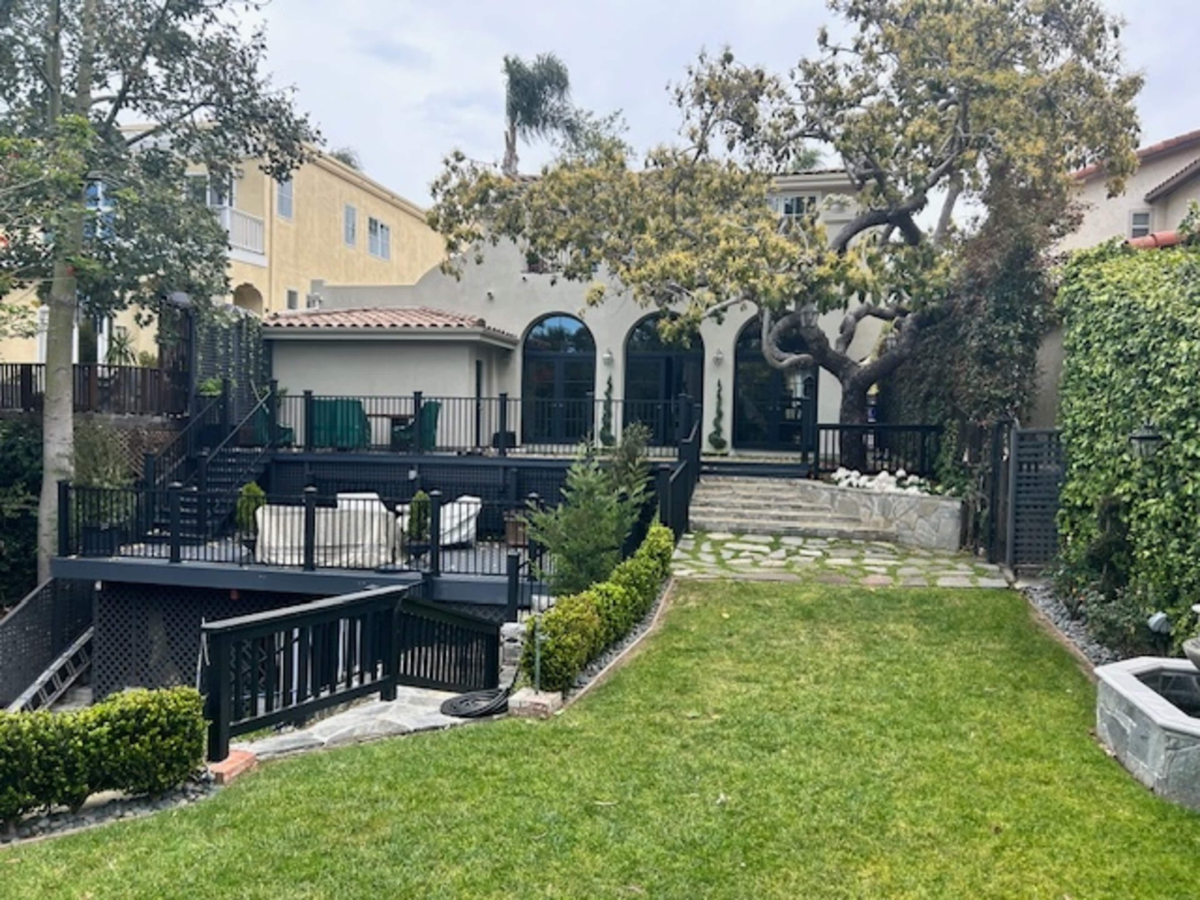 A landscaped backyard featuring a stone pathway, lush green grass, and a multi-level deck surrounded by trees and shrubbery.