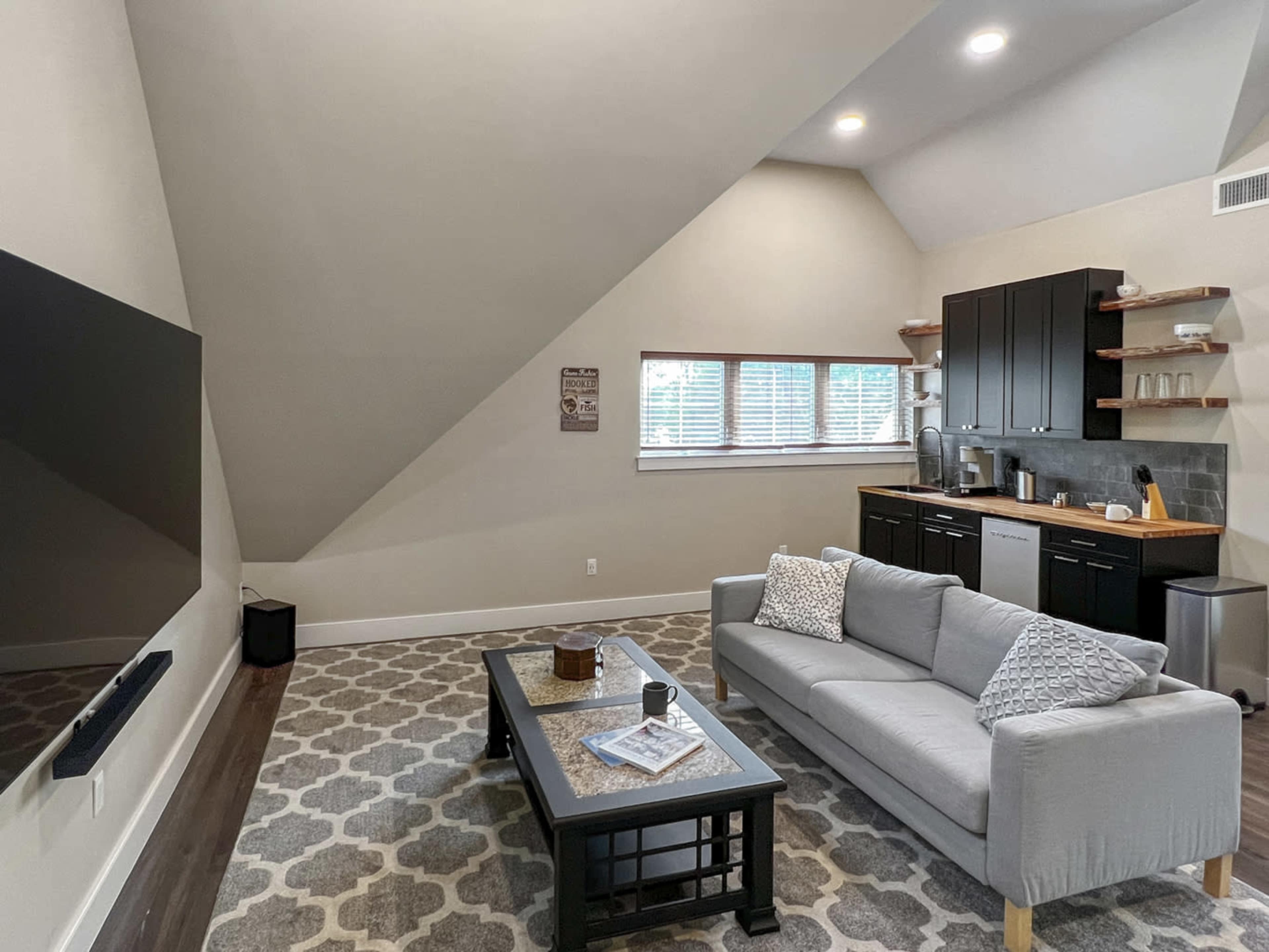 A modern living area with a sloped ceiling, featuring a gray sofa, a coffee table, and a small kitchenette with dark cabinetry.