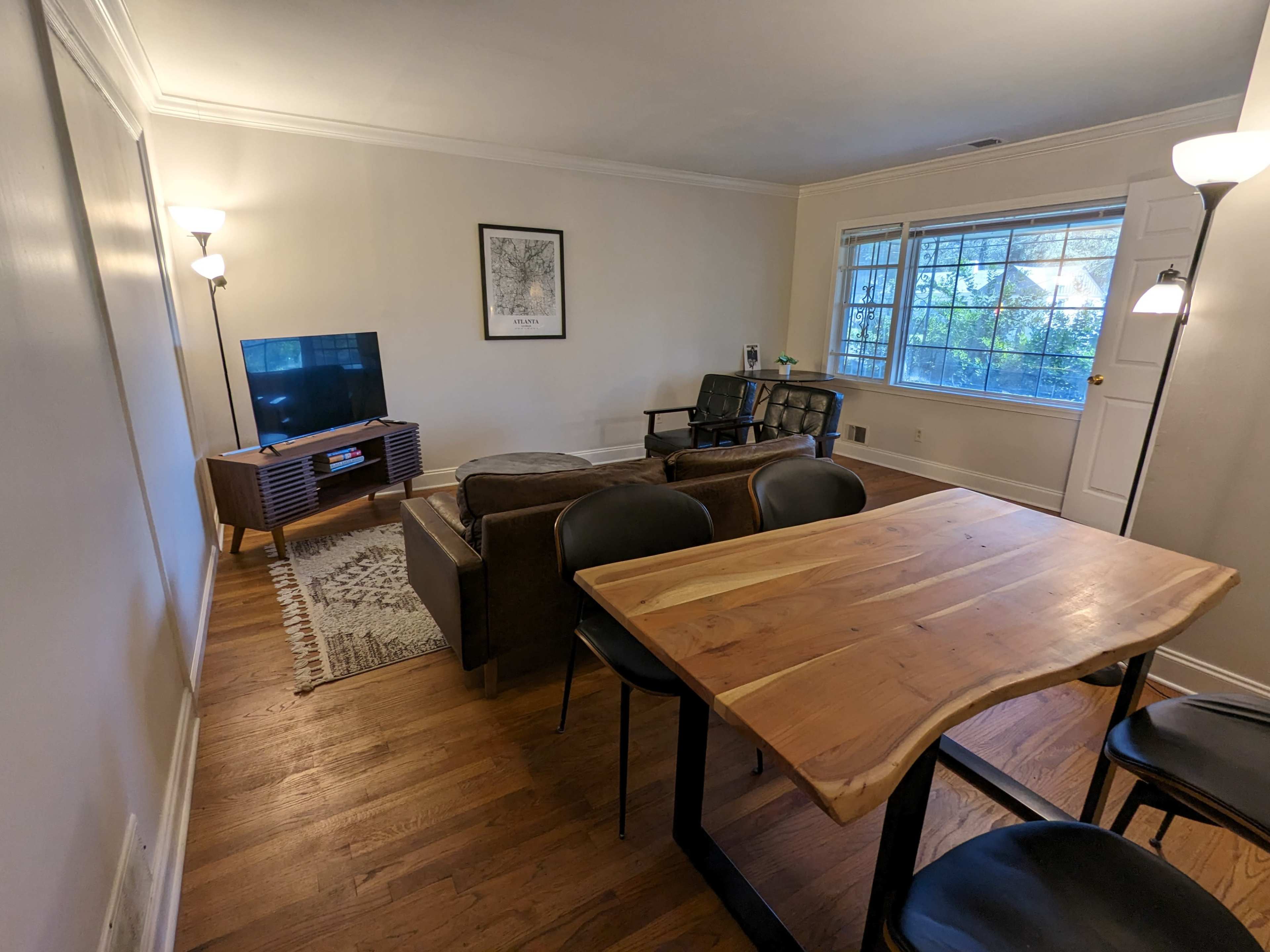 The image shows a living room with a seating area, a television on a stand, a dining table, and large windows allowing natural light.
