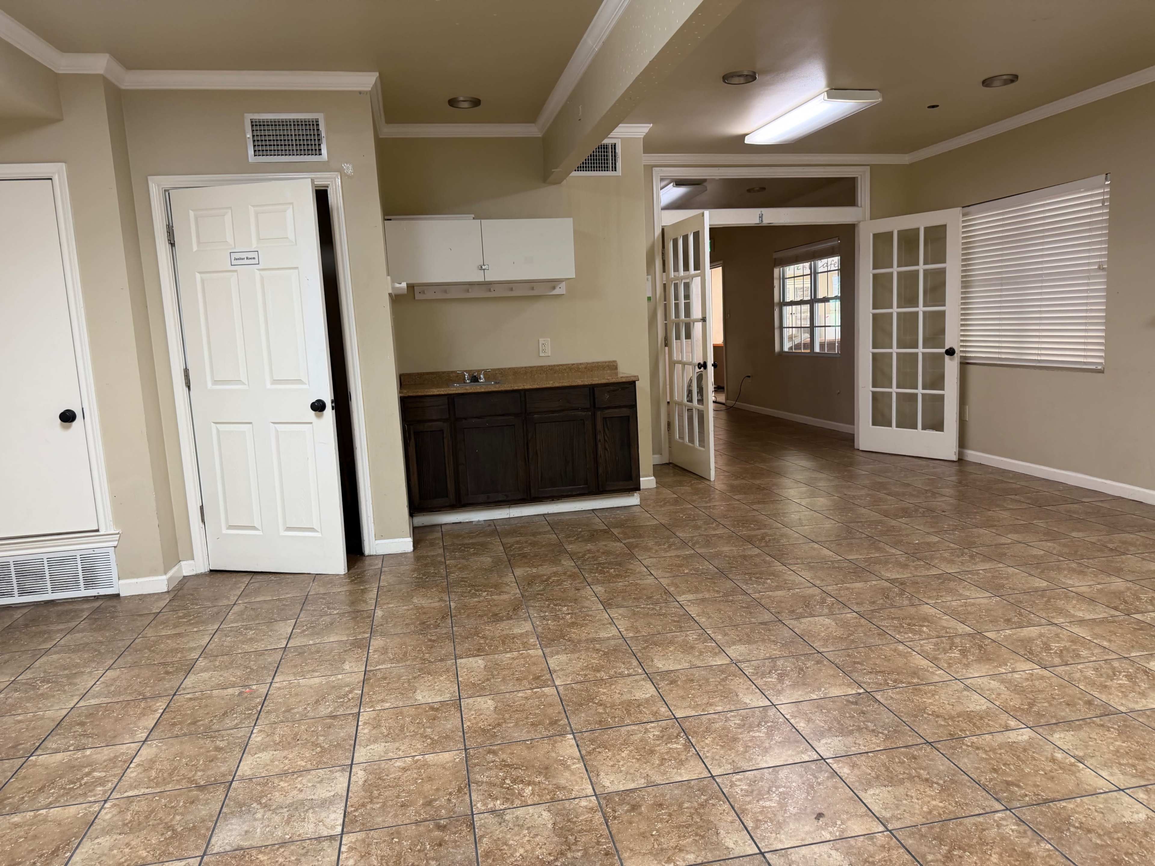 The image shows a spacious, tiled room with an open layout, featuring a kitchenette area and a pair of French doors leading to another room.