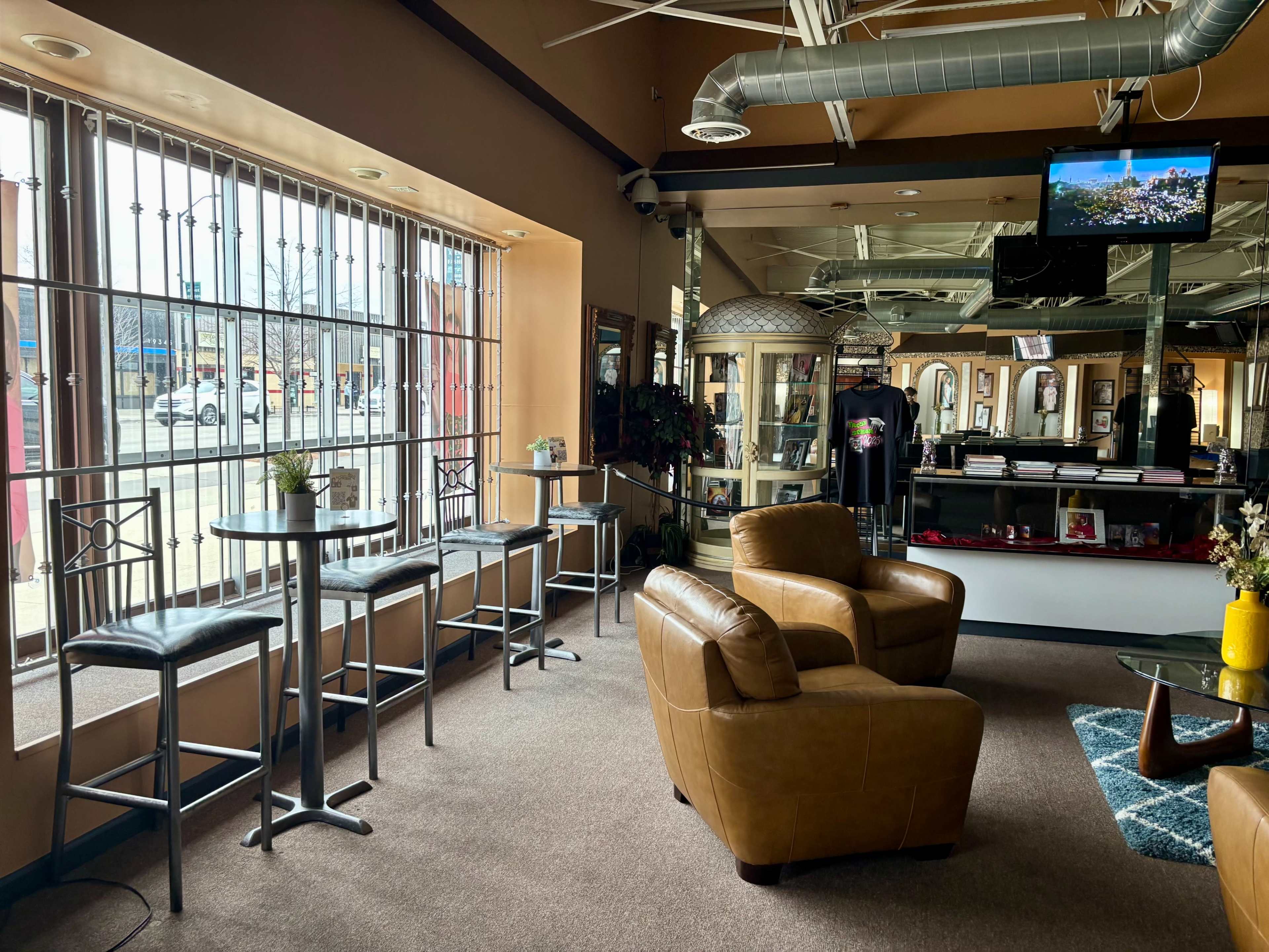 The image shows a cozy lounge area with two leather chairs and several tall tables near large, barred windows letting in natural light.