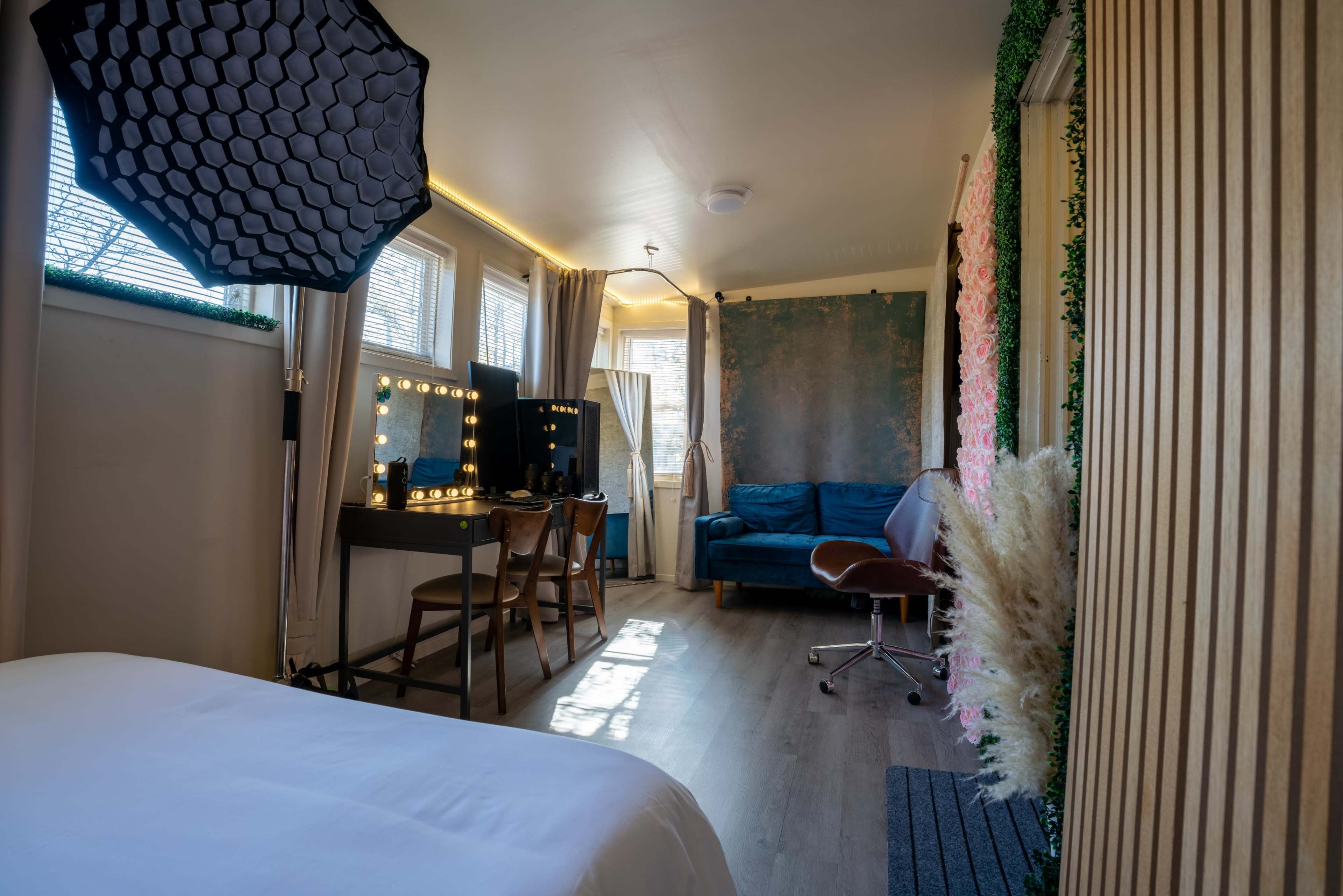 A well-lit room featuring a bed in the foreground, a makeup station with lights, a blue sofa, and a textured wall.