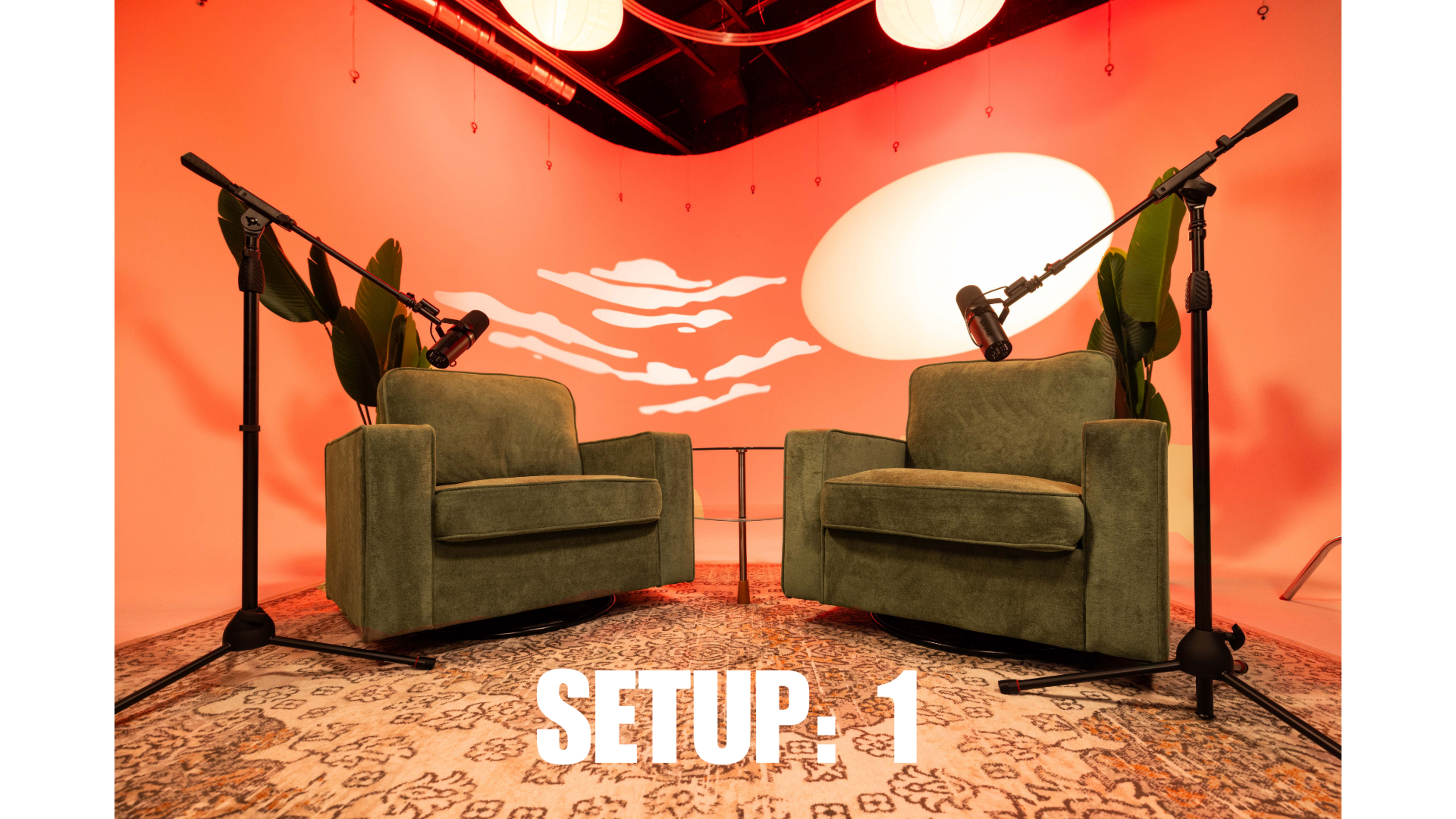 The image shows a modern interview setup featuring two green chairs with microphones on stands positioned in front of an orange backdrop with a white abstract design.