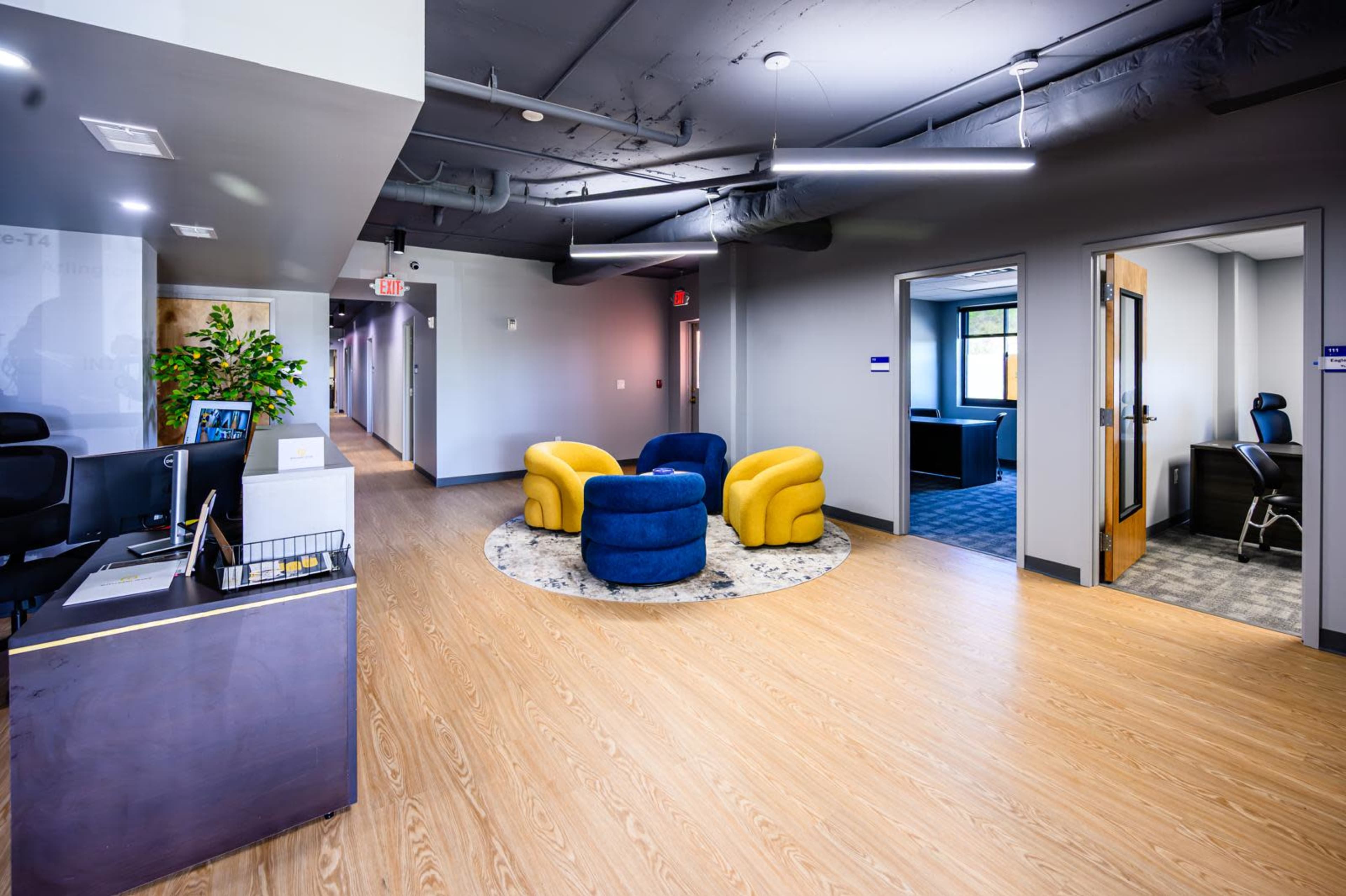 The image shows a modern office lobby featuring a reception desk, two yellow and blue chairs arranged around a circular rug, and a corridor leading to additional office spaces.