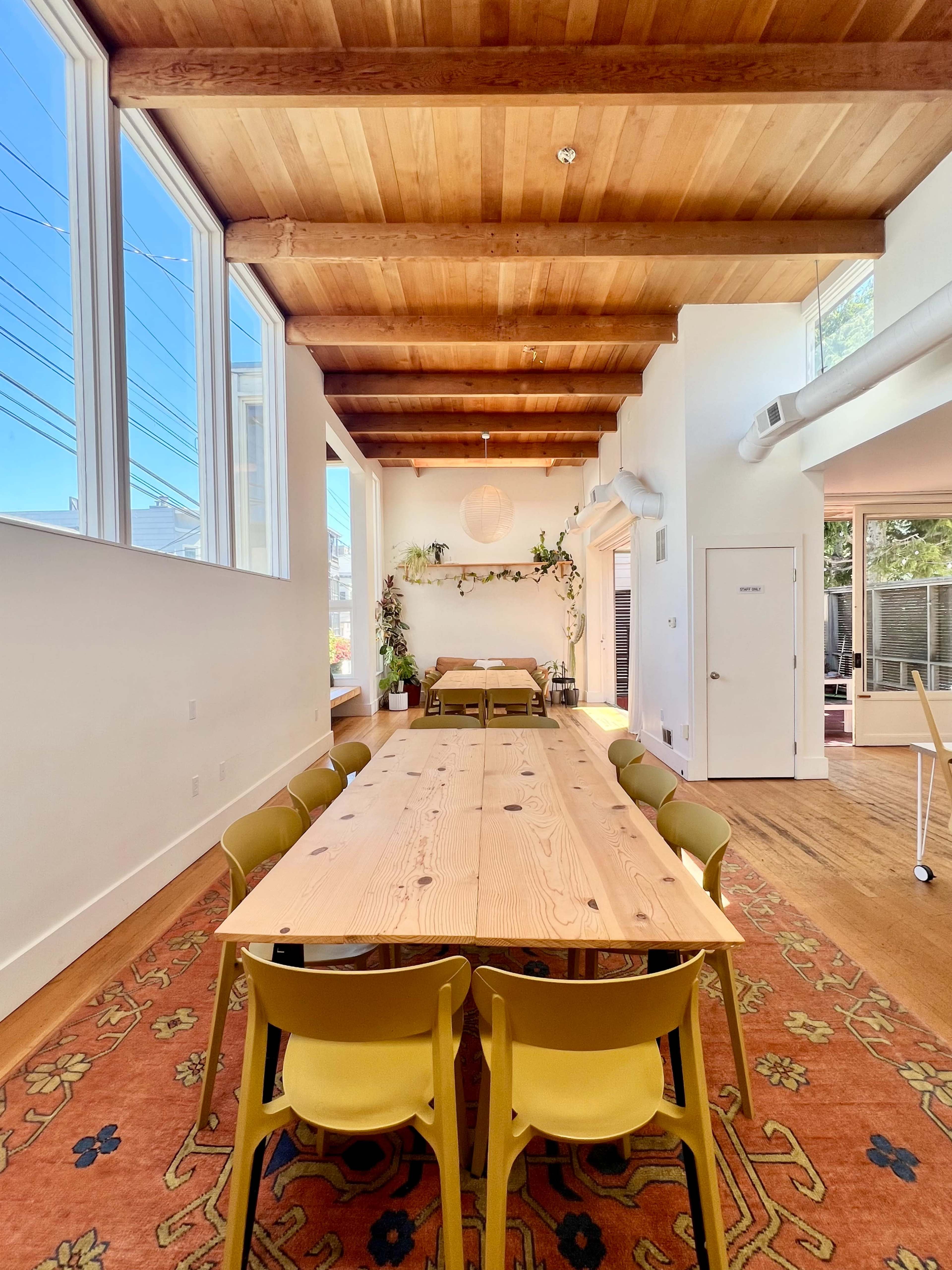 The image shows a well-lit room featuring a long wooden dining table surrounded by yellow chairs, with wooden beams on the ceiling and large windows allowing natural light in.