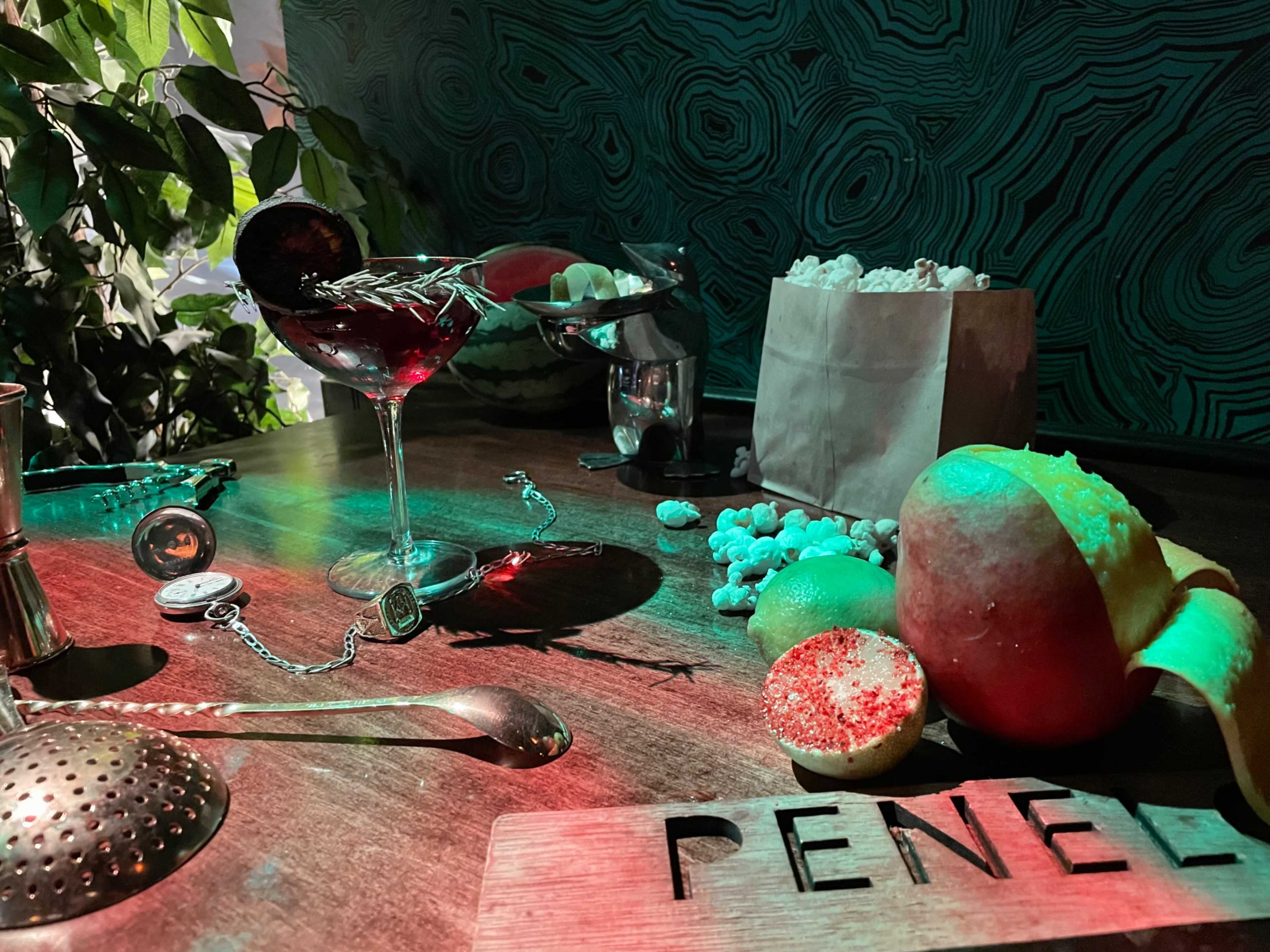 The scene features a table with a cocktail glass filled with a dark drink, popcorn, an apple, and a wooden sign that reads "PENEL."