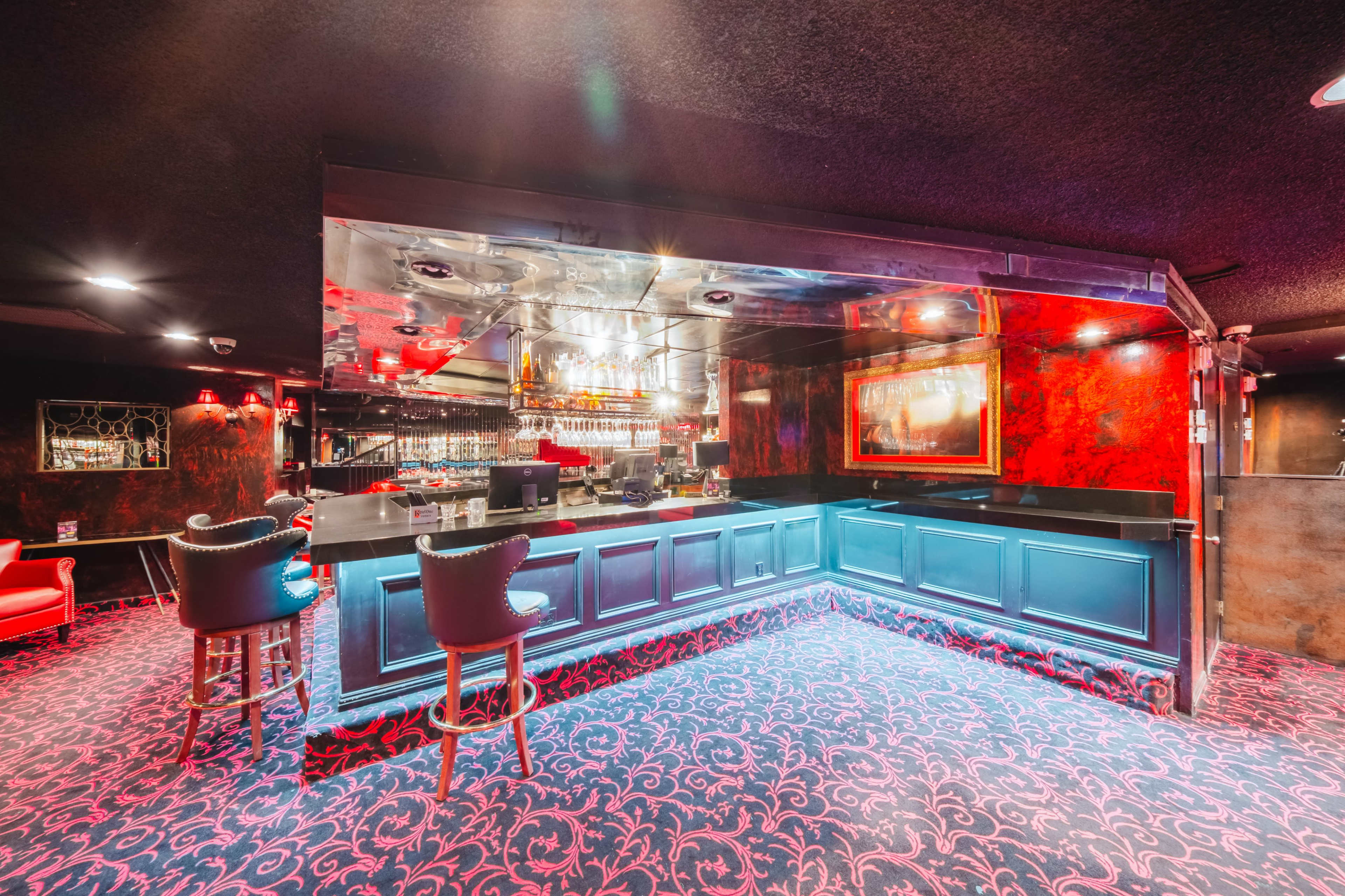 The image shows a dimly lit bar with a sleek counter, illuminated by blue lights, and surrounded by stylish seating in a vibrant red and black decor.