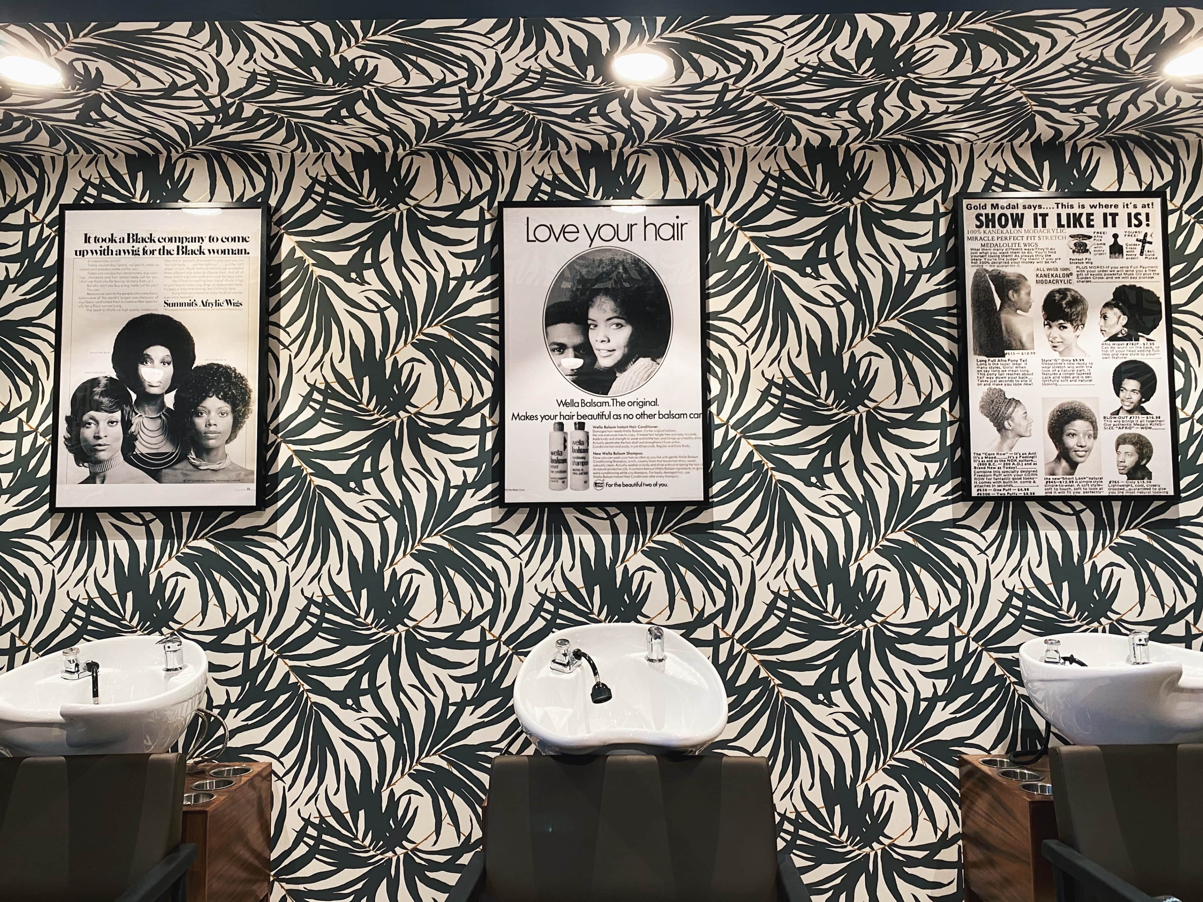 A salon interior featuring three framed posters on a decorative leaf-patterned wall, along with two hair-washing stations that have white basins.