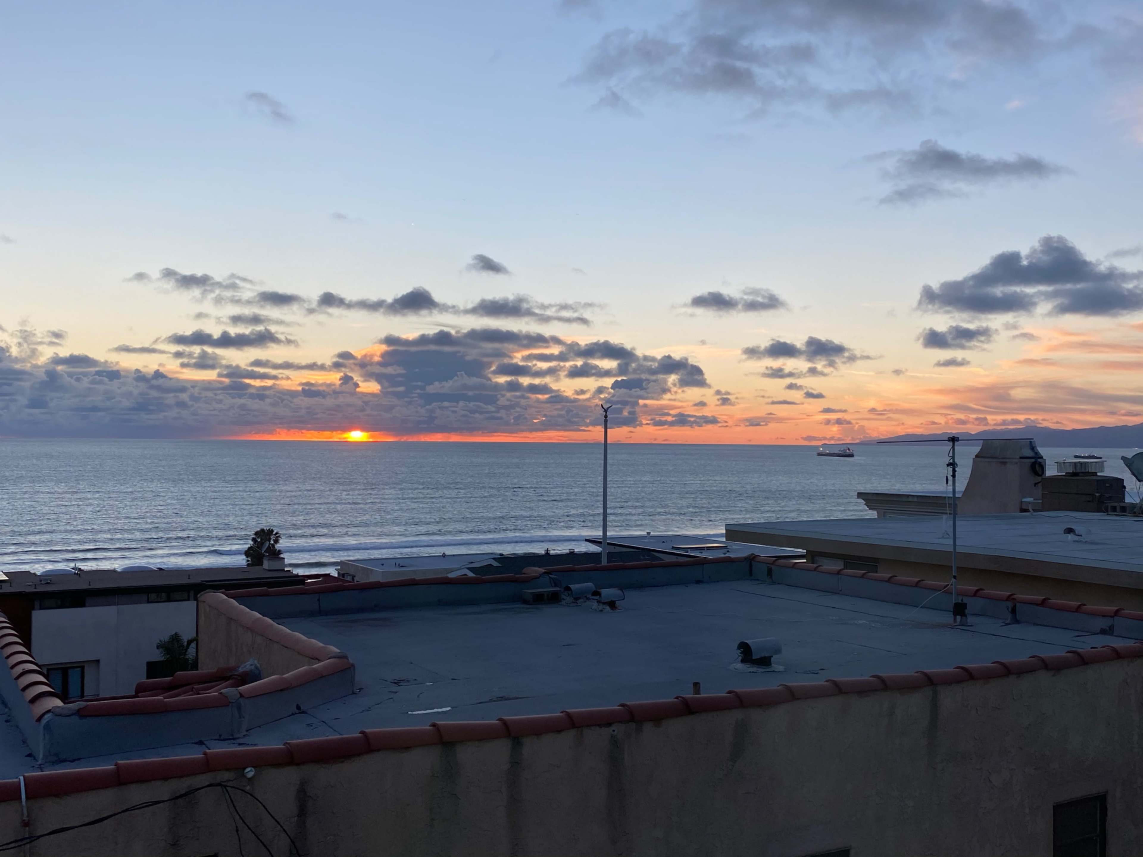 The sun sets over the ocean, casting hues of orange and yellow across the horizon, while clouds linger above.