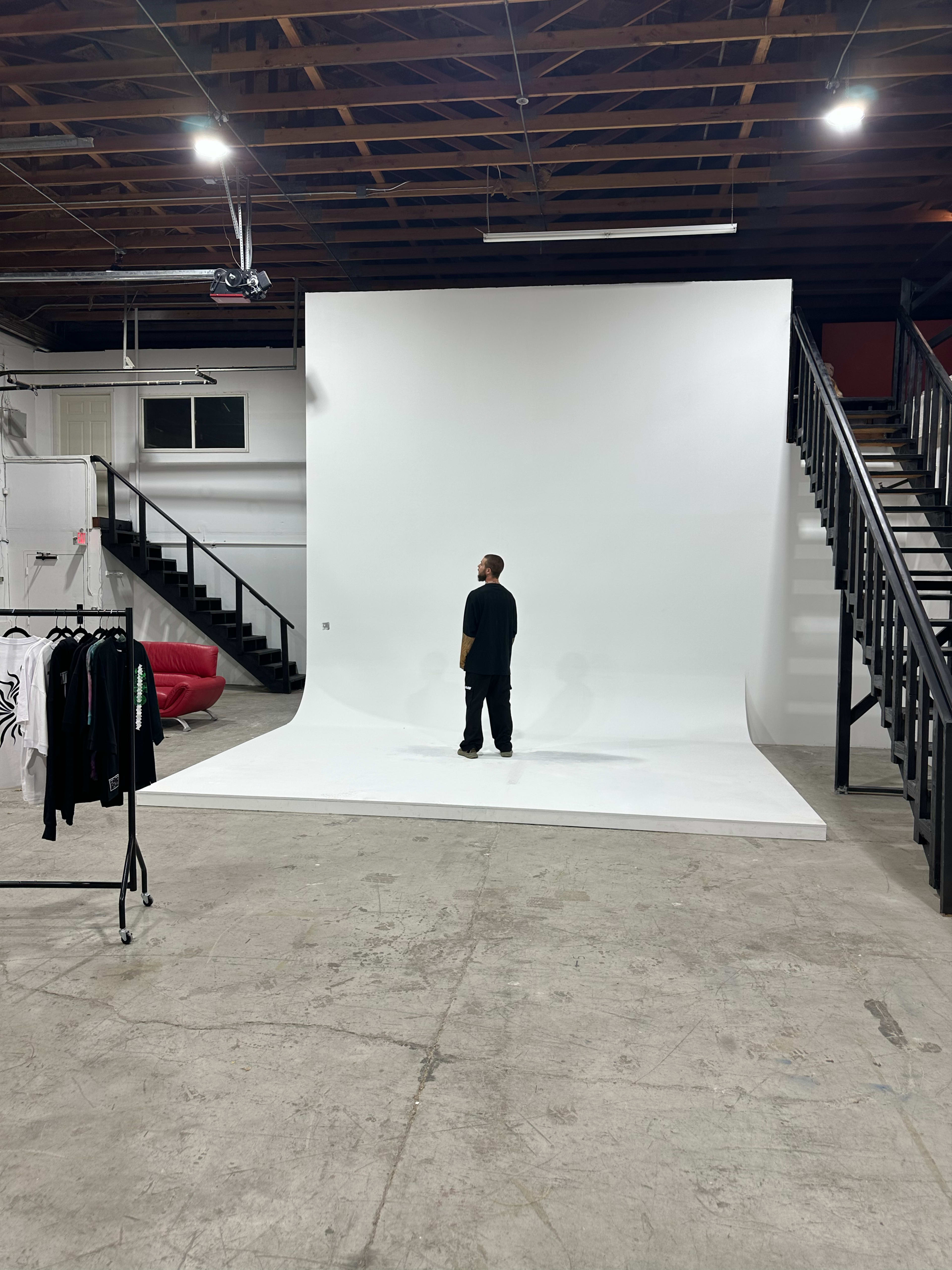 A person stands alone in a spacious studio with a large white backdrop and a staircase in the background.