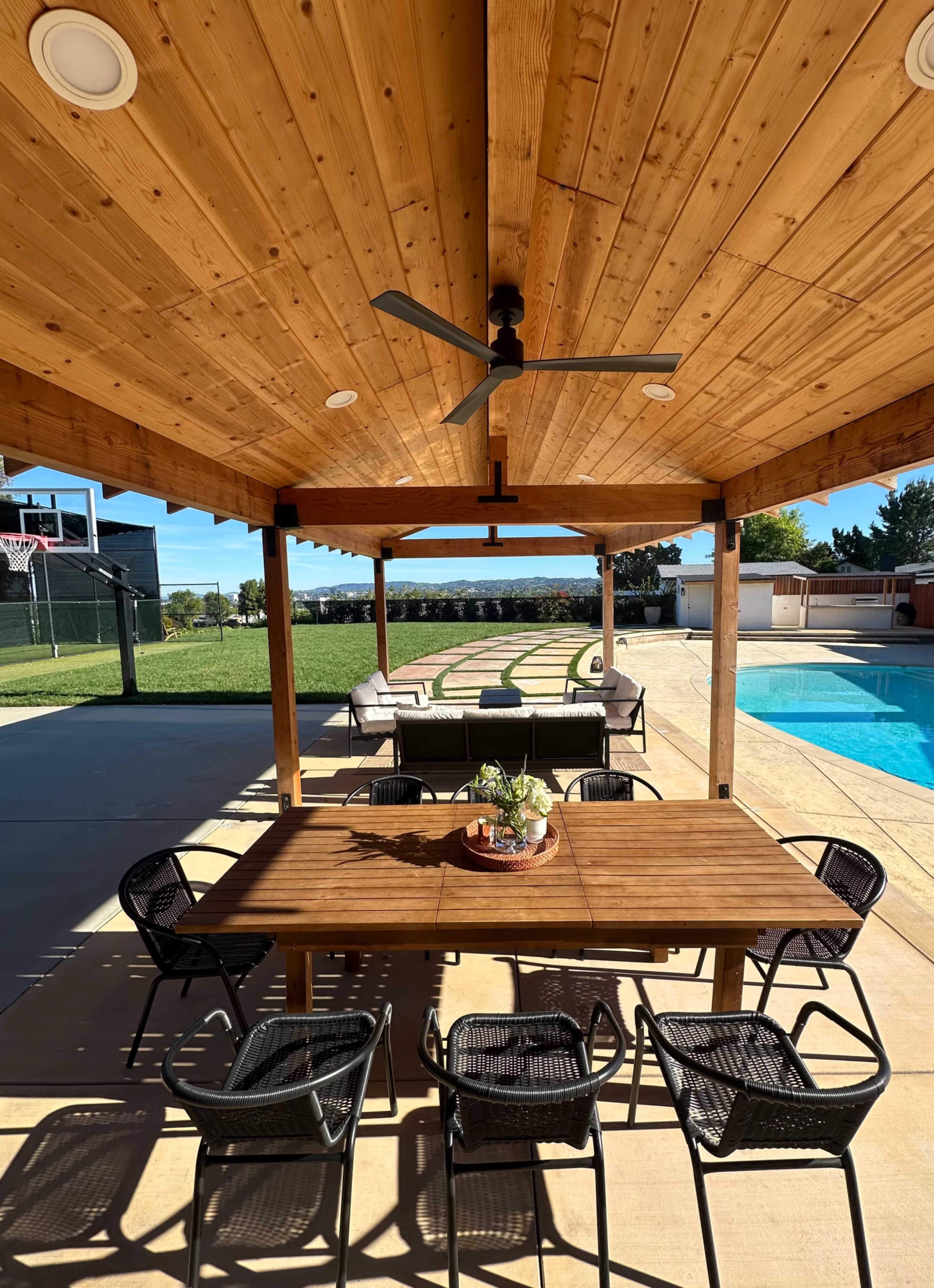 A covered patio features a wooden table with eight black chairs overlooking a pool area and a grassy yard.
