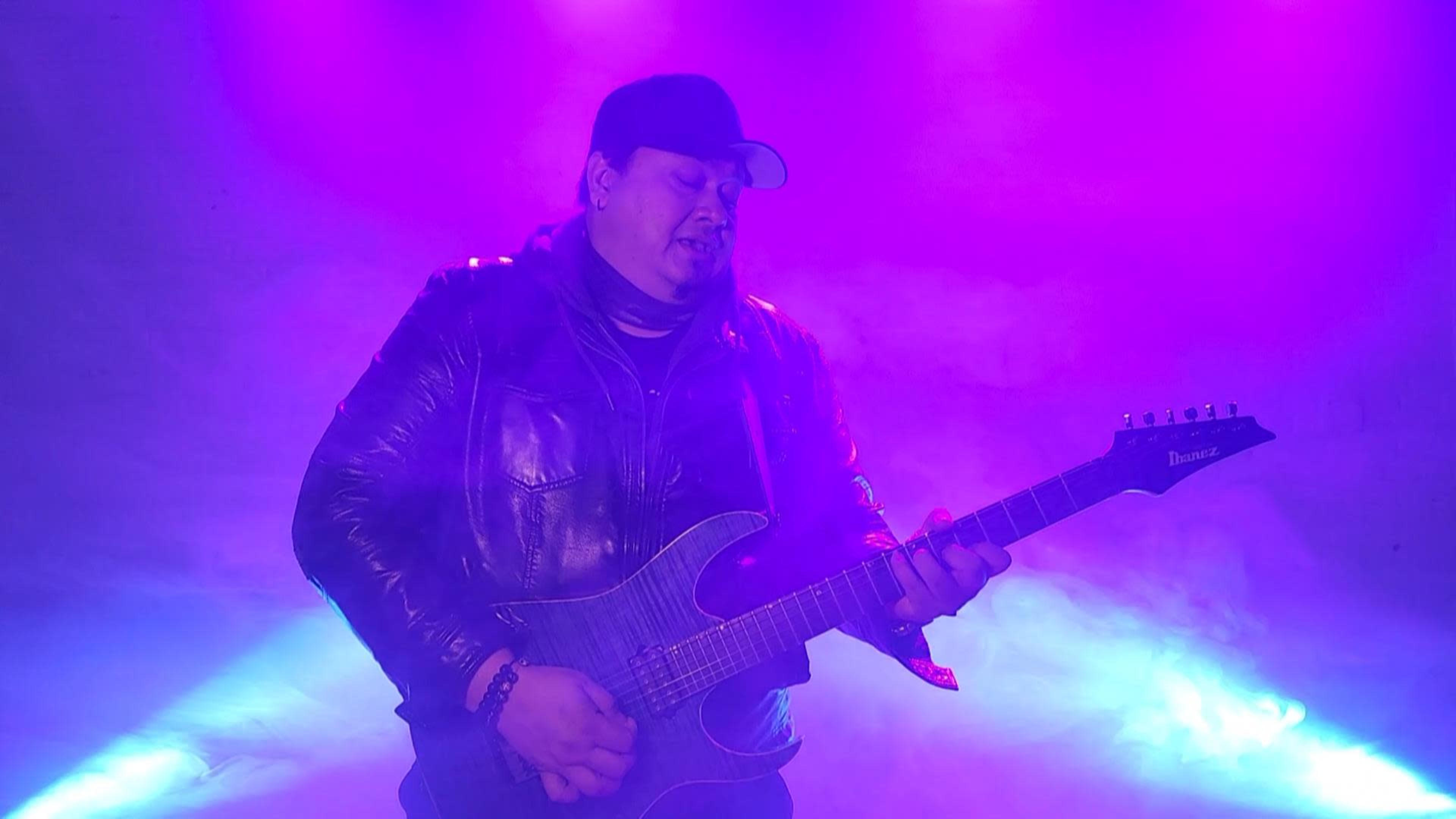 A musician wearing a leather jacket and cap plays an electric guitar under purple and blue stage lights.