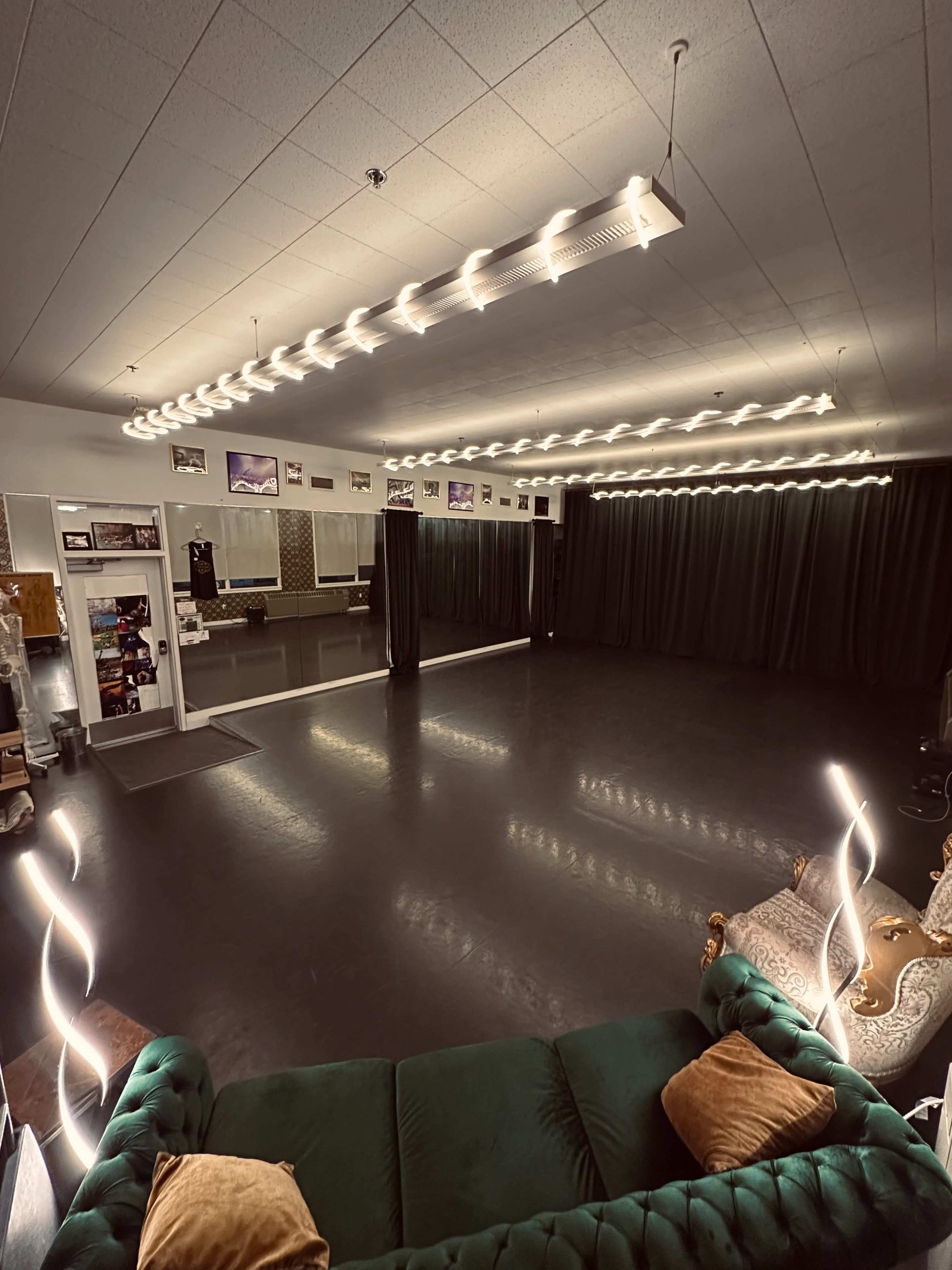 The image shows a spacious dance studio with dark flooring, mirrored walls, and ambient lighting, featuring a green sofa and decorative pillows in one corner.