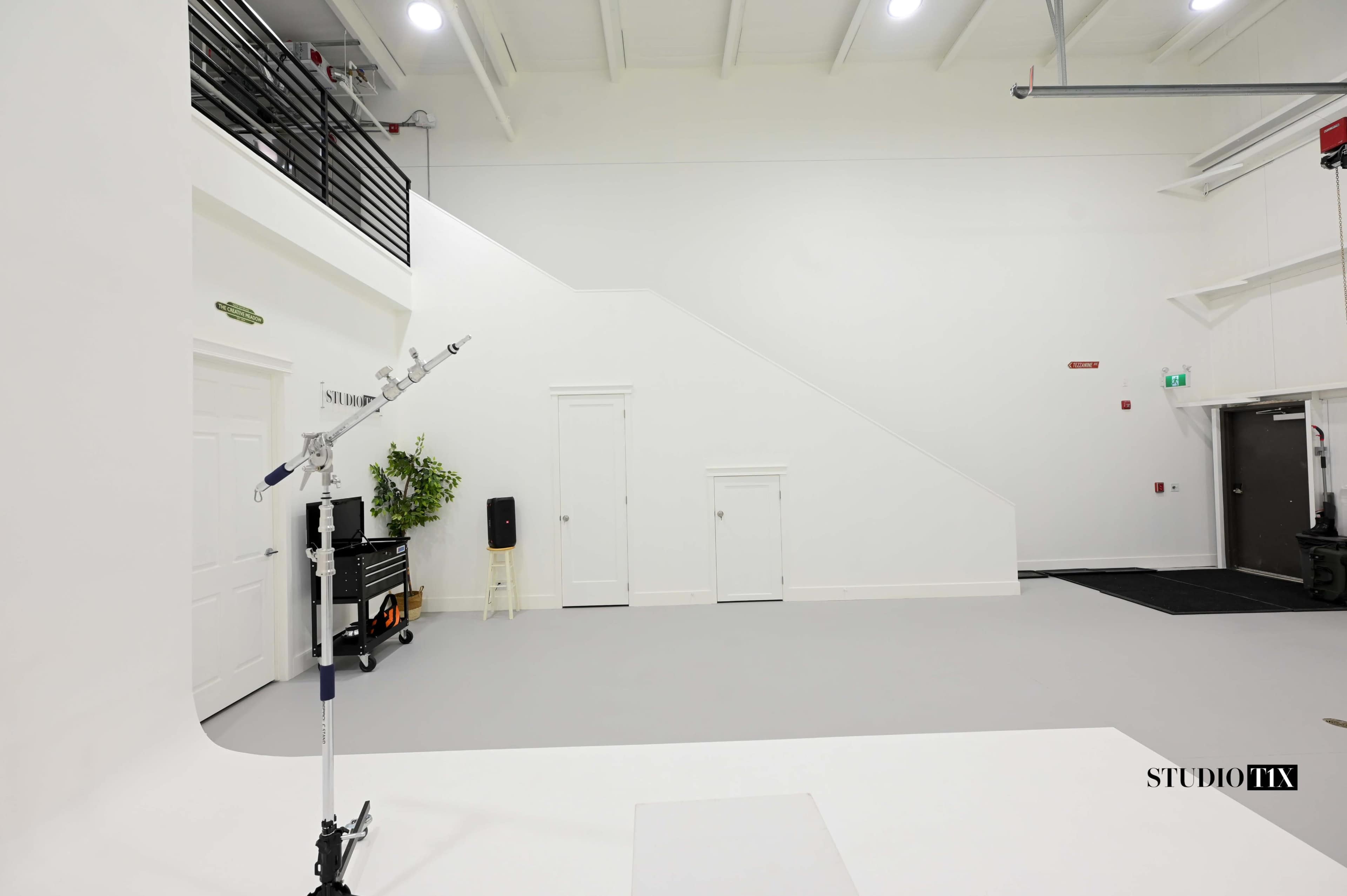 The image shows a spacious, well-lit studio with white walls, a gray floor, and a staircase leading to a second level.