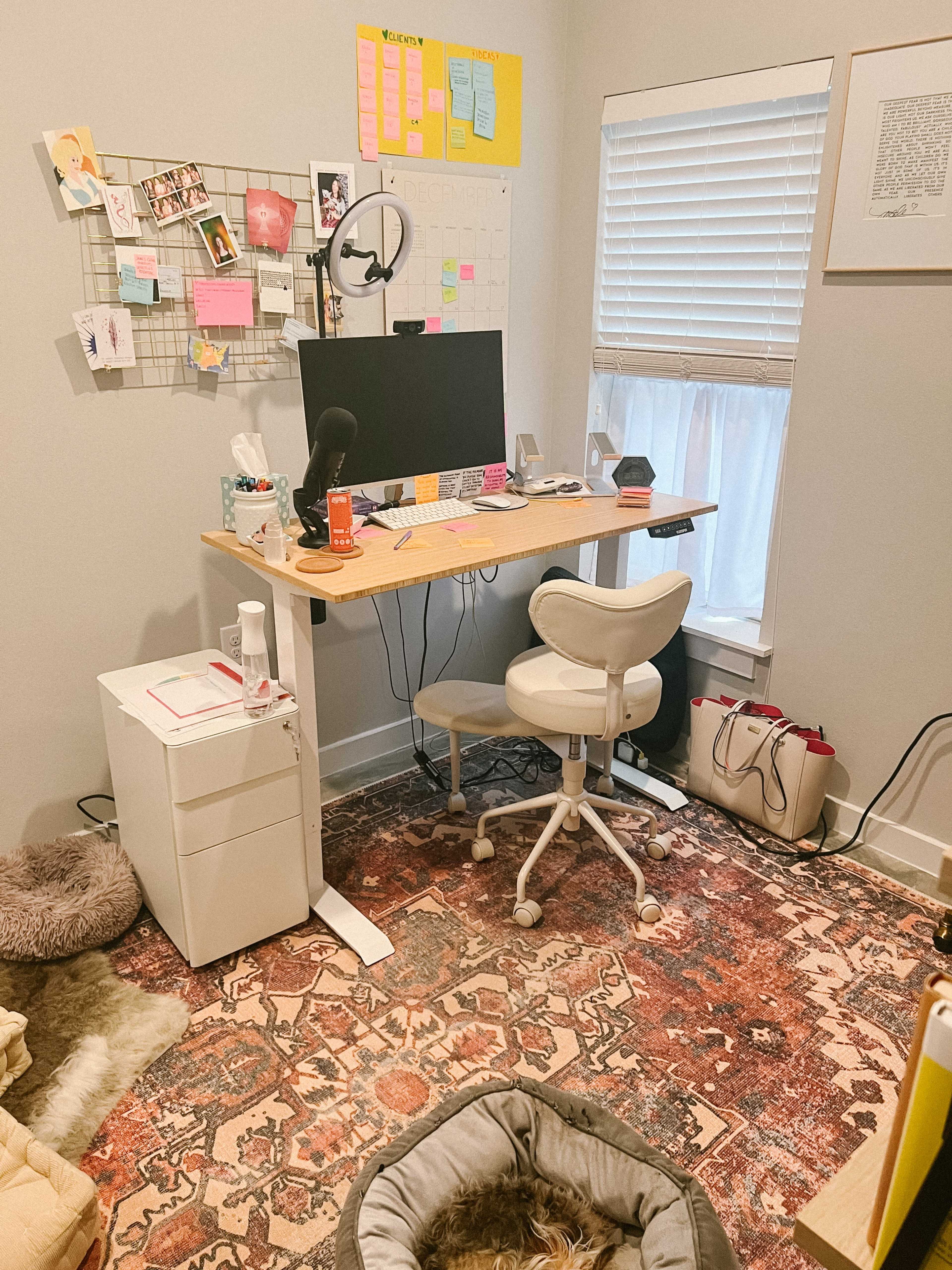 A home office features a desk with a computer monitor, a chair, various office supplies, and a rug, alongside a wall with a grid displaying photos and notes.