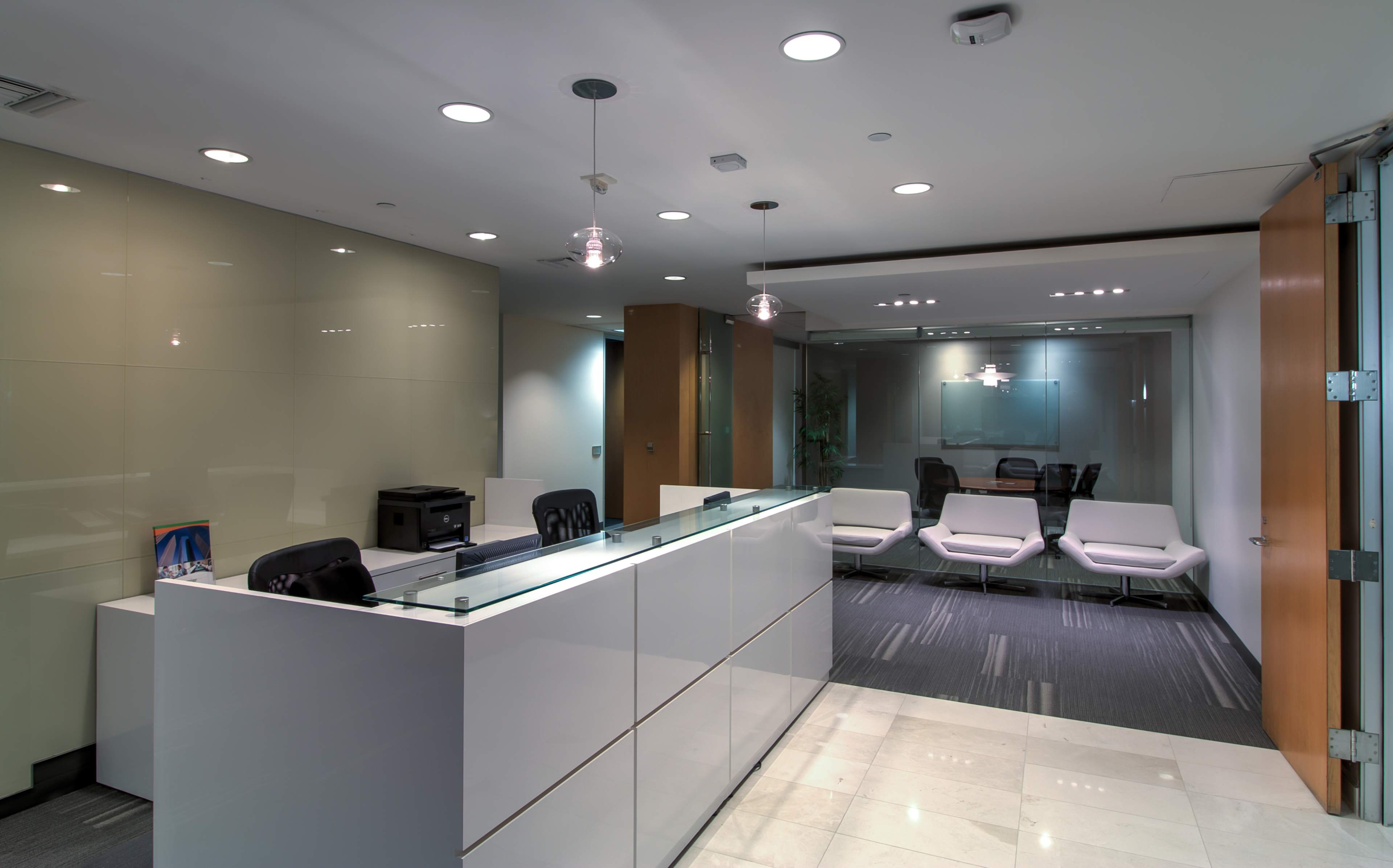 A modern office reception area with a white counter, chairs, and glass partitions separating it from a conference room.