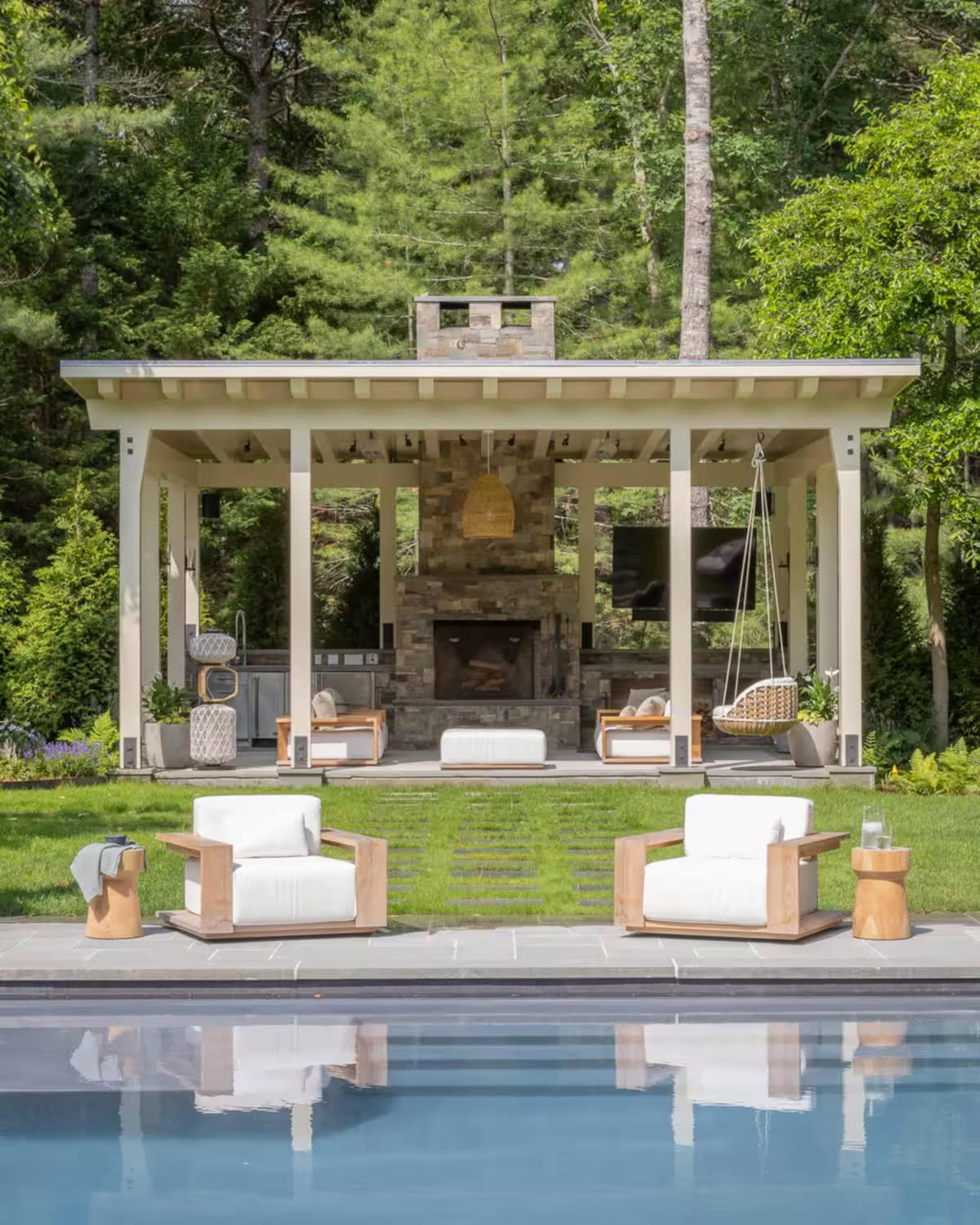 A modern outdoor living area features a stone fireplace, an adjacent kitchen, and comfortable seating arranged around a swimming pool.