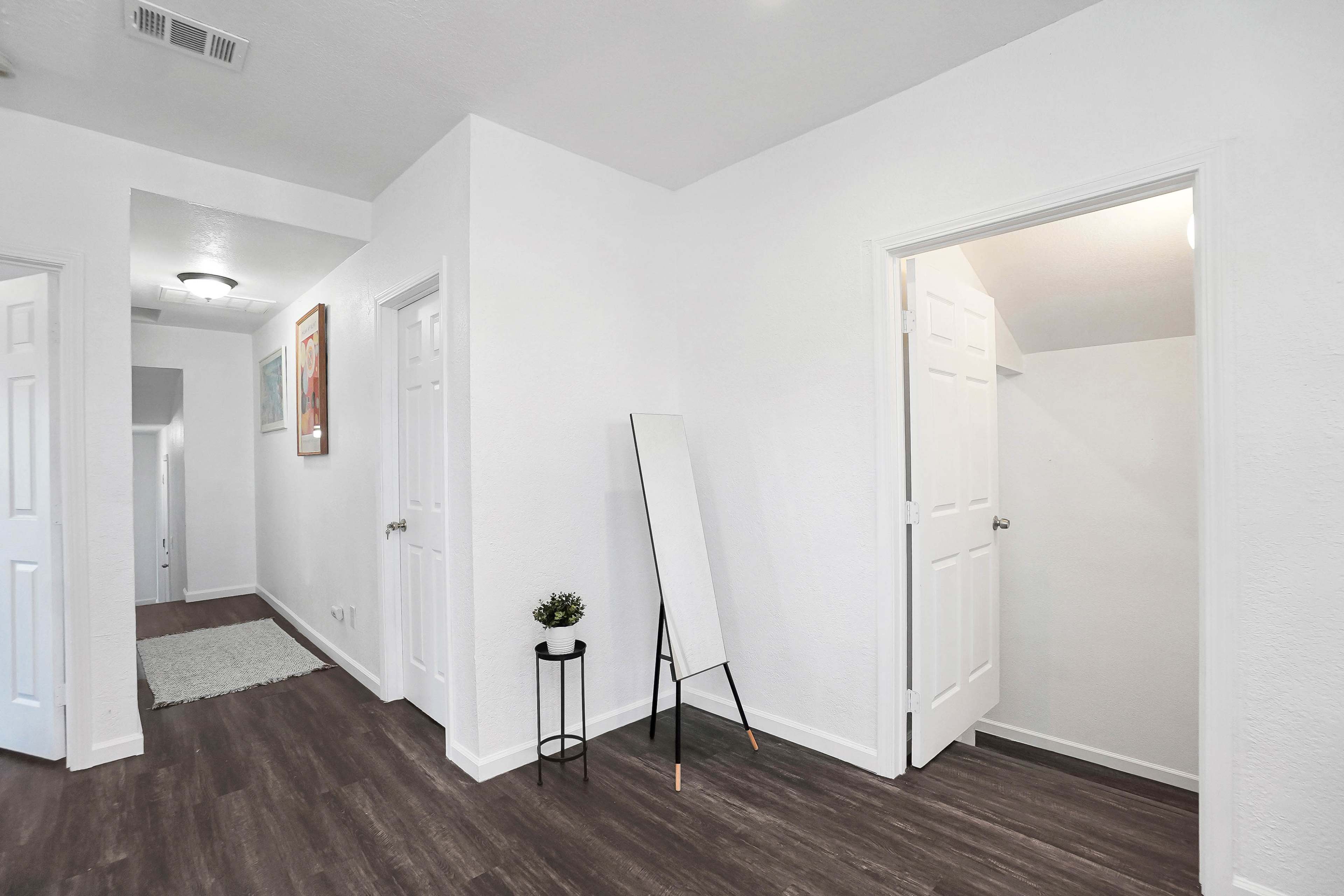 The image shows a well-lit hallway with white walls, hardwood flooring, and a standing mirror next to a small table.