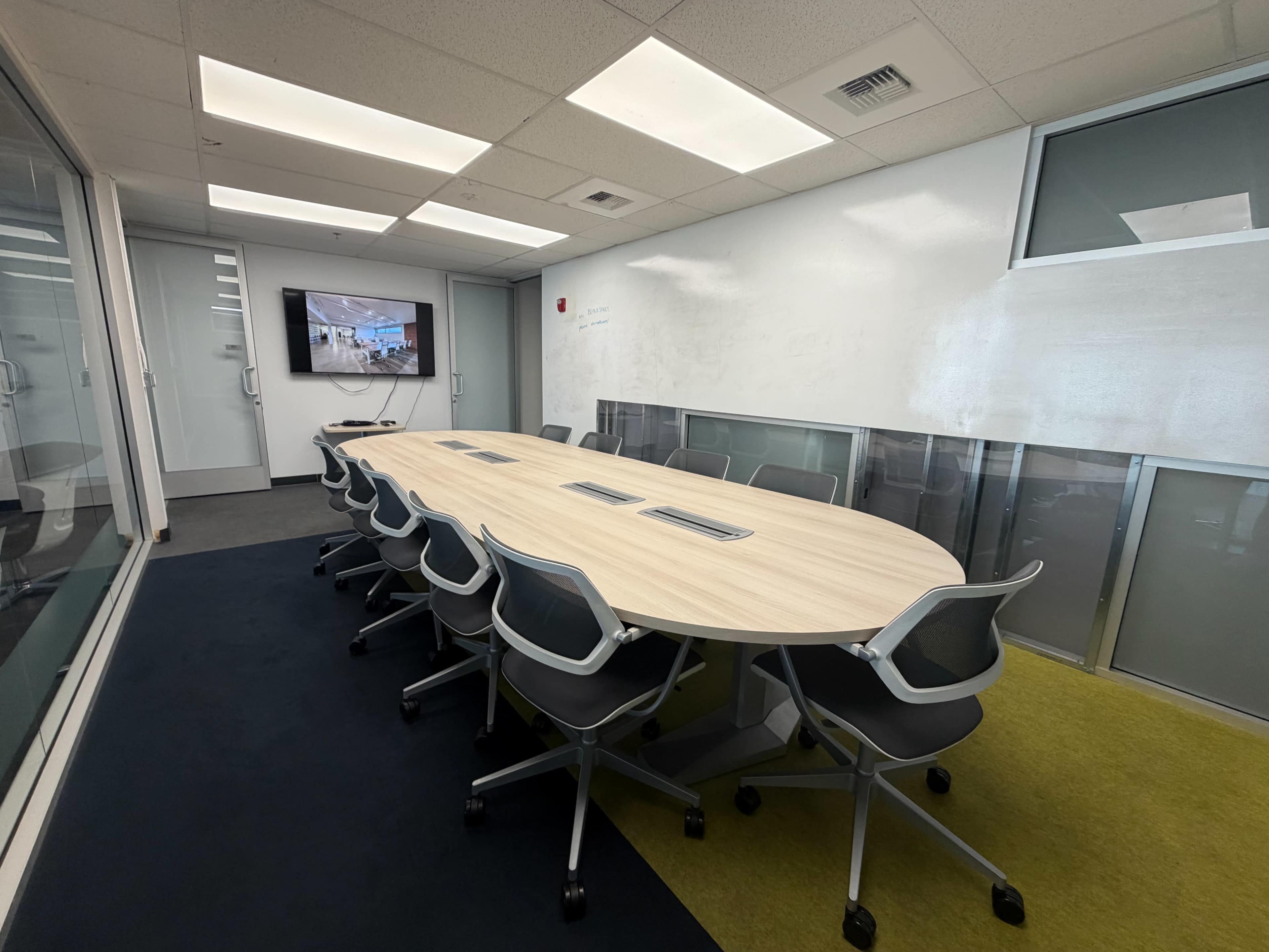 The image shows a modern conference room with a large oval table surrounded by chairs, a television mounted on the wall, and glass partitions.