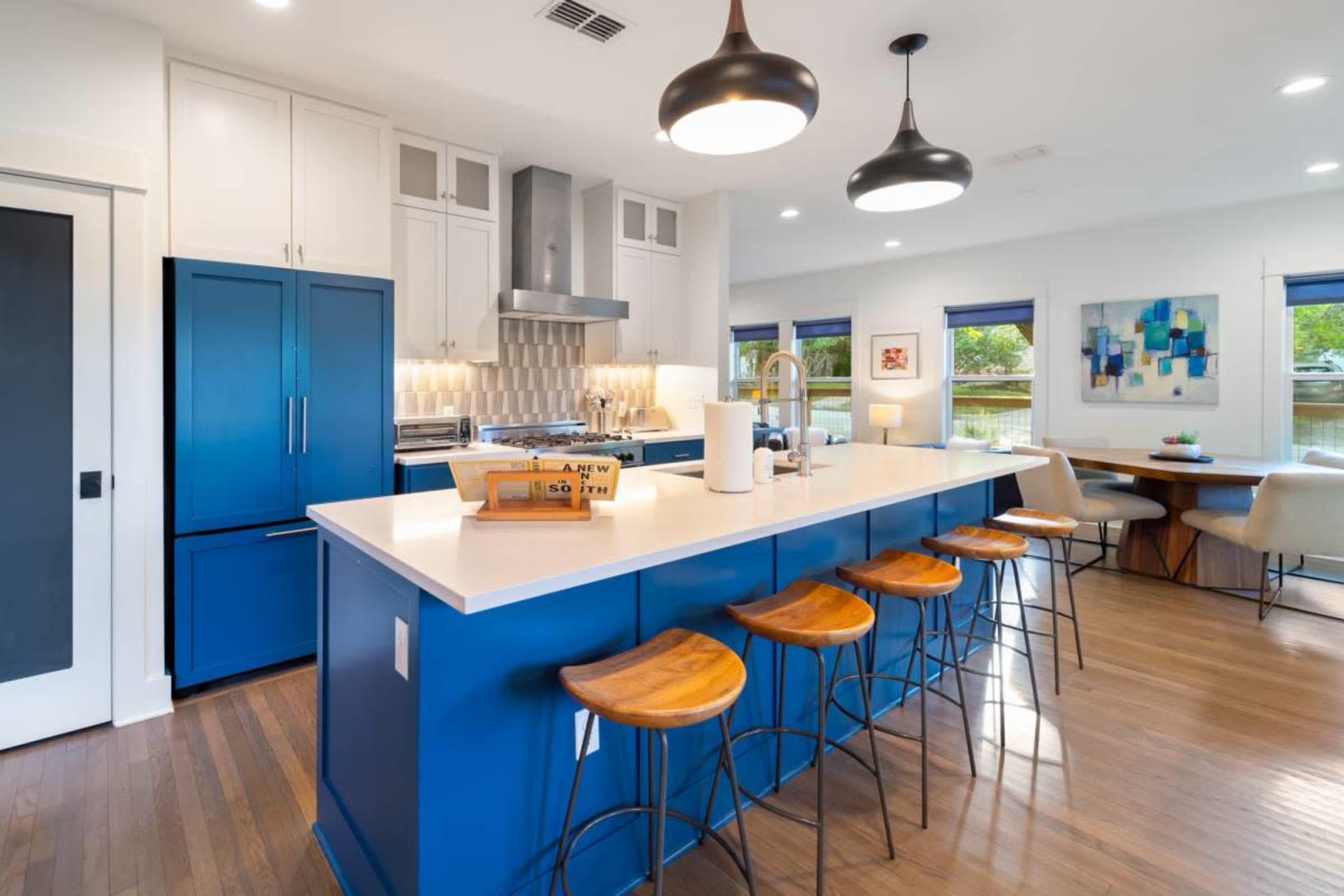 A modern kitchen features a blue island with wooden bar stools, stainless steel appliances, and ample natural light from windows.