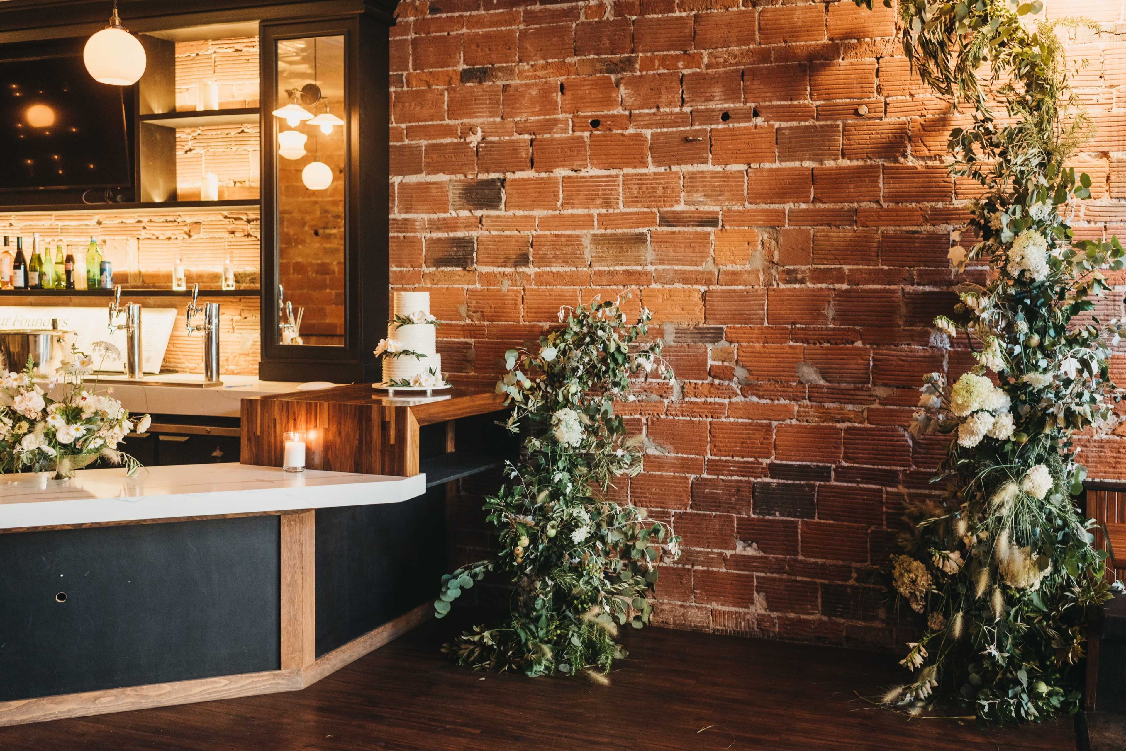 A decorated interior featuring a brick wall with greenery and floral arrangements near a bar area.