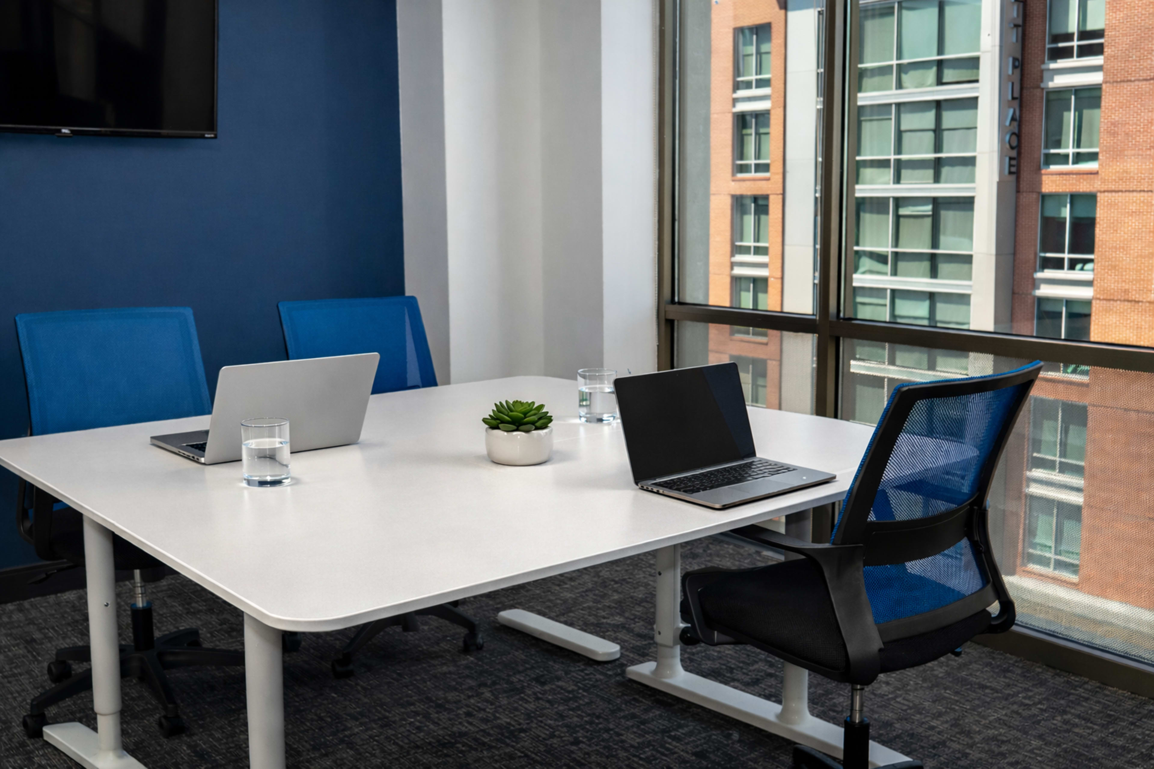 A modern conference room features a white table with three laptops, two glasses of water, and a small plant, surrounded by blue chairs and large windows displaying an urban view.