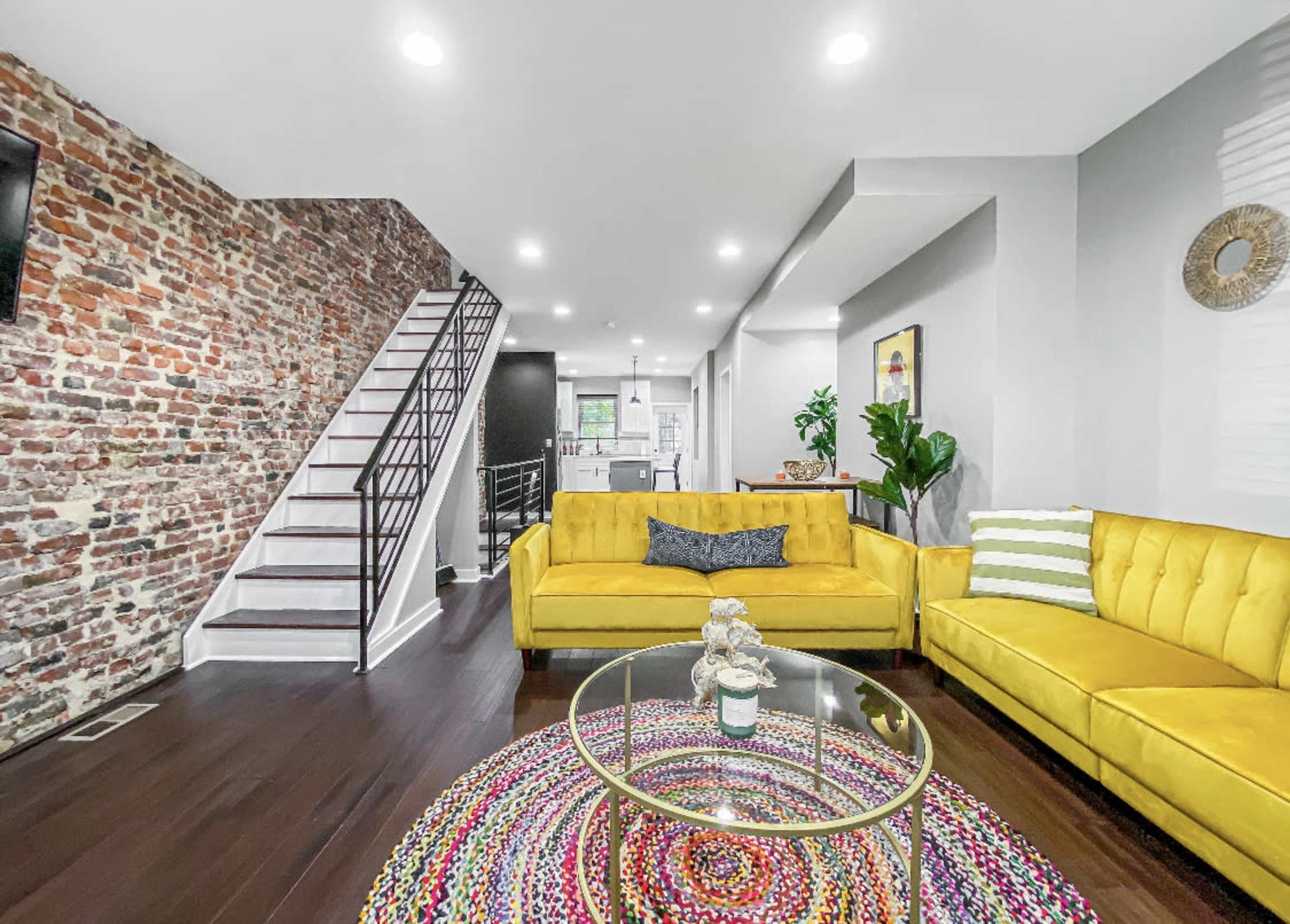 The image shows a modern living room with a yellow couch, a round colorful rug, exposed brick walls, and a staircase leading to the upper level.