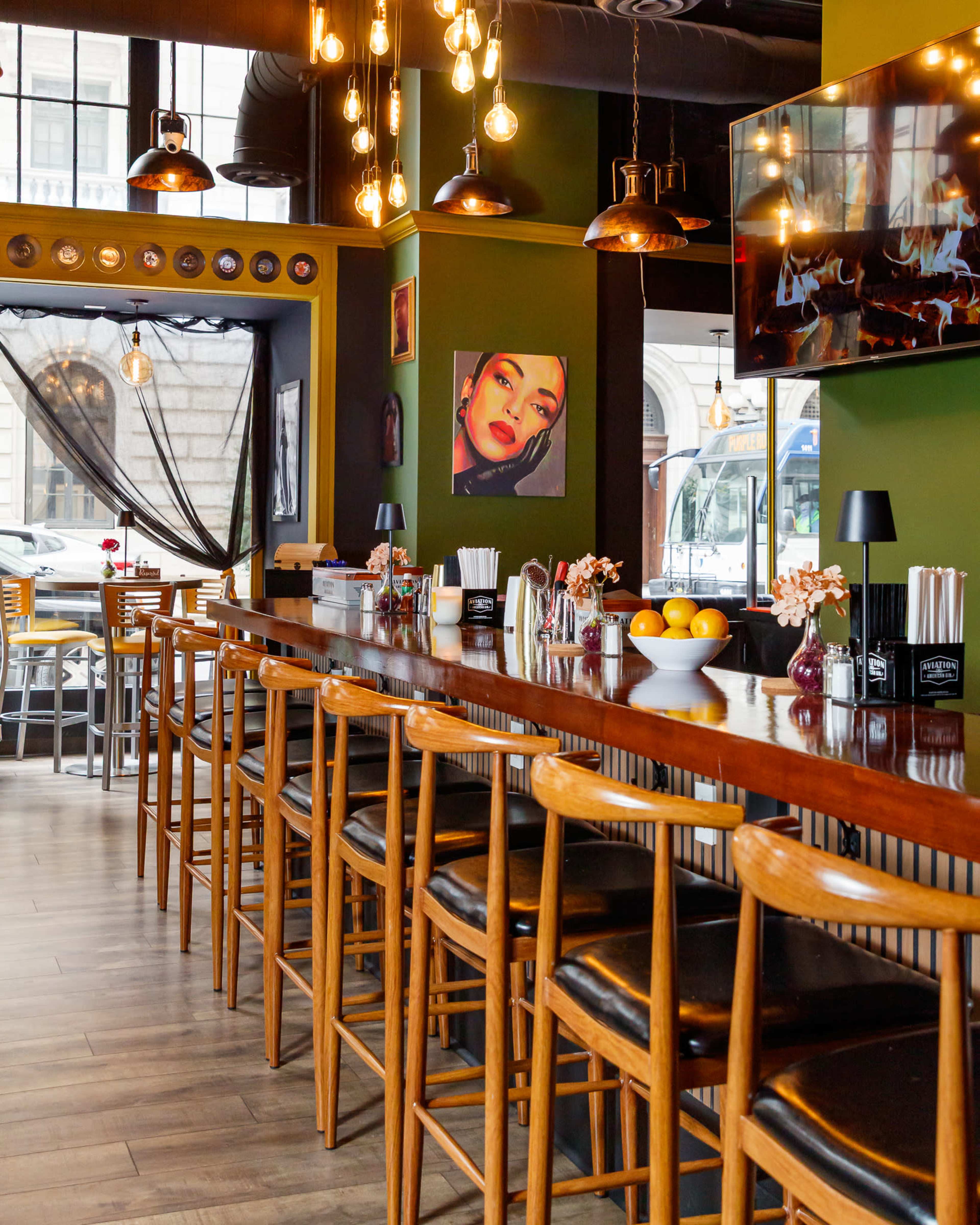 The image shows a modern bar area with wooden stools, a long polished countertop, and decorative lighting, set in a vibrant green-walled space featuring artwork and a television.