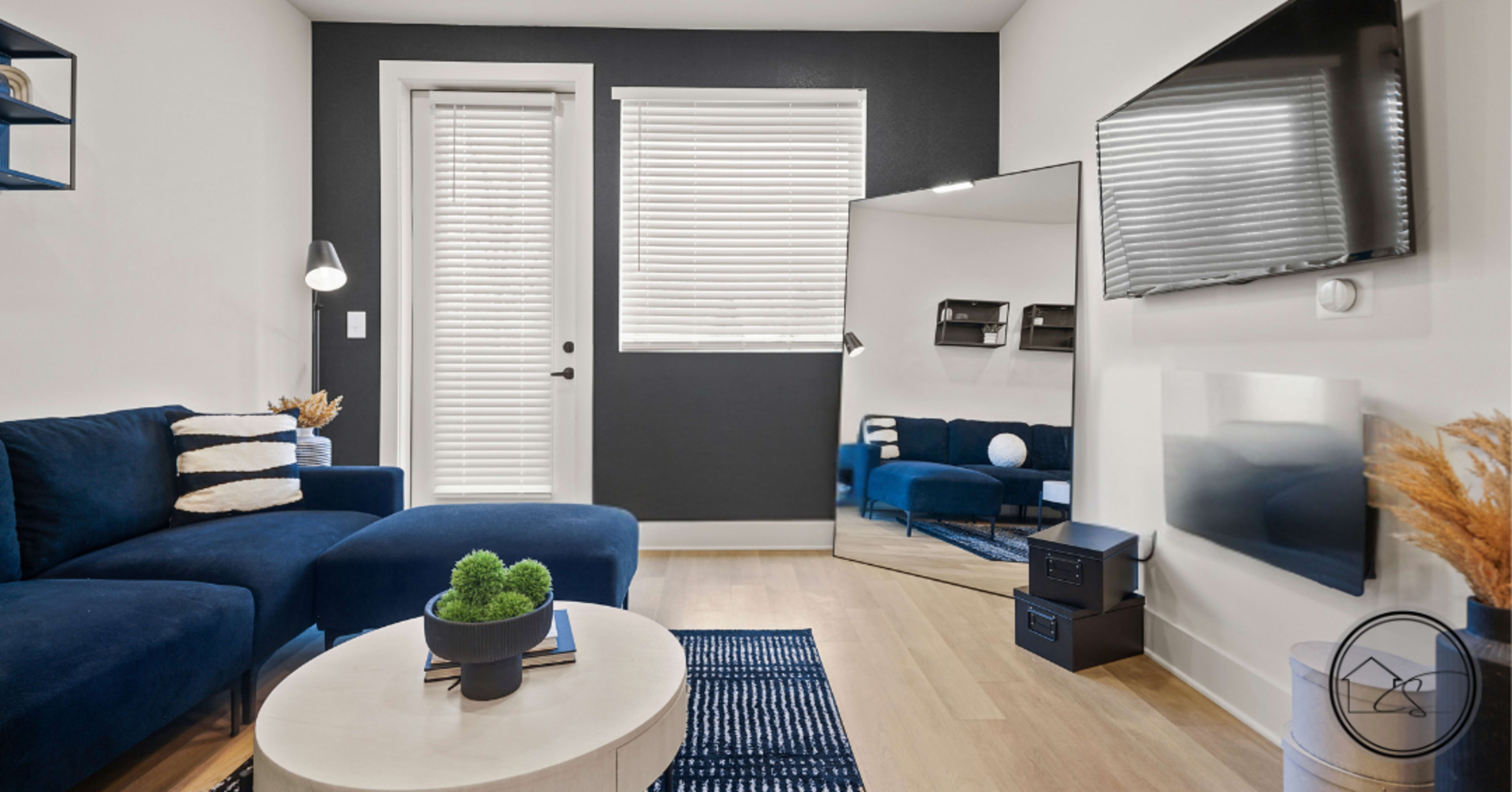 The image depicts a modern living room with a blue sectional sofa, a round coffee table, and a large wall-mounted mirror beside a television.