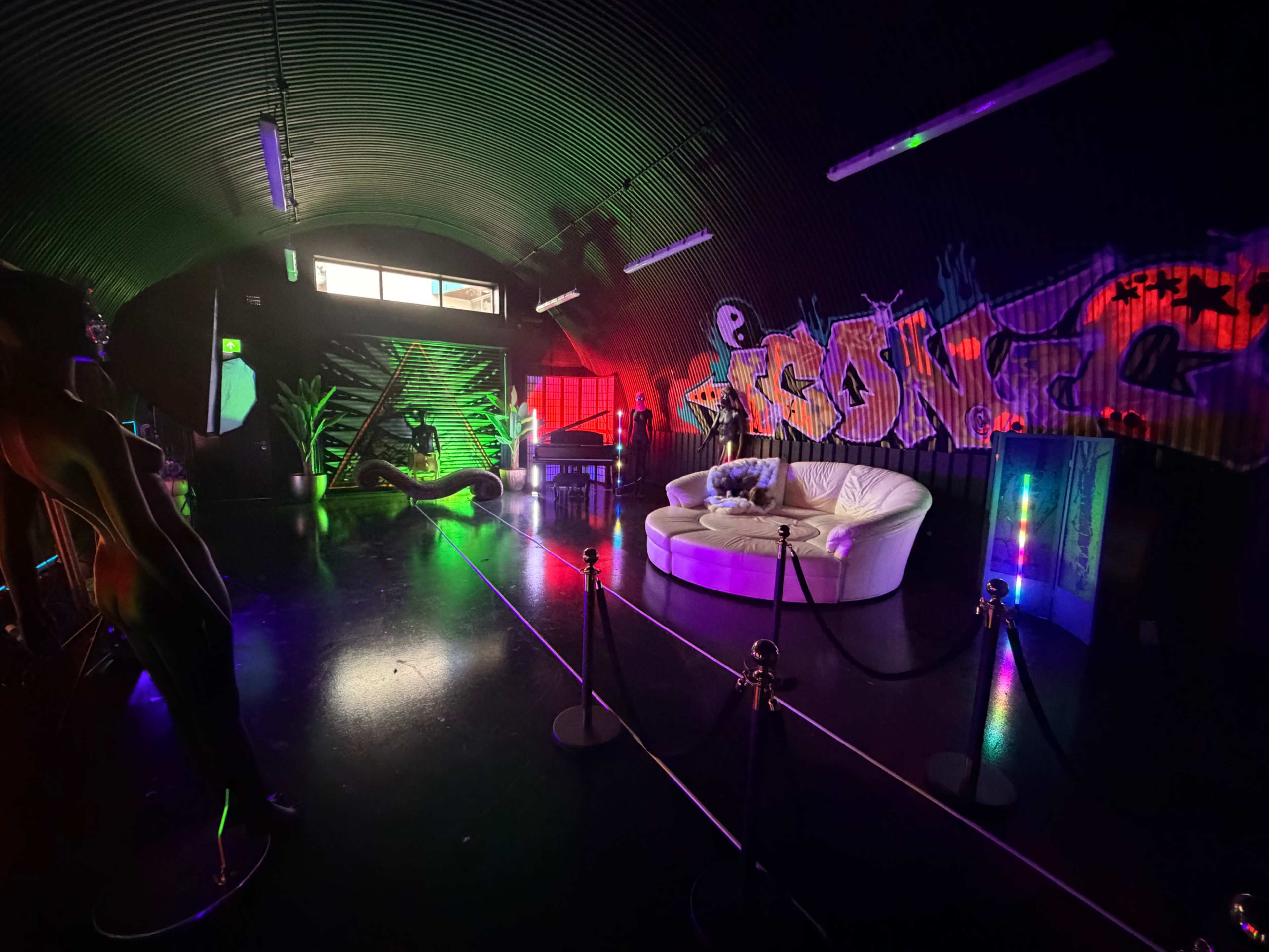 The image depicts a dimly lit, stylized lounge area featuring a white circular couch, colorful lighting, and graffiti decorations on the walls.