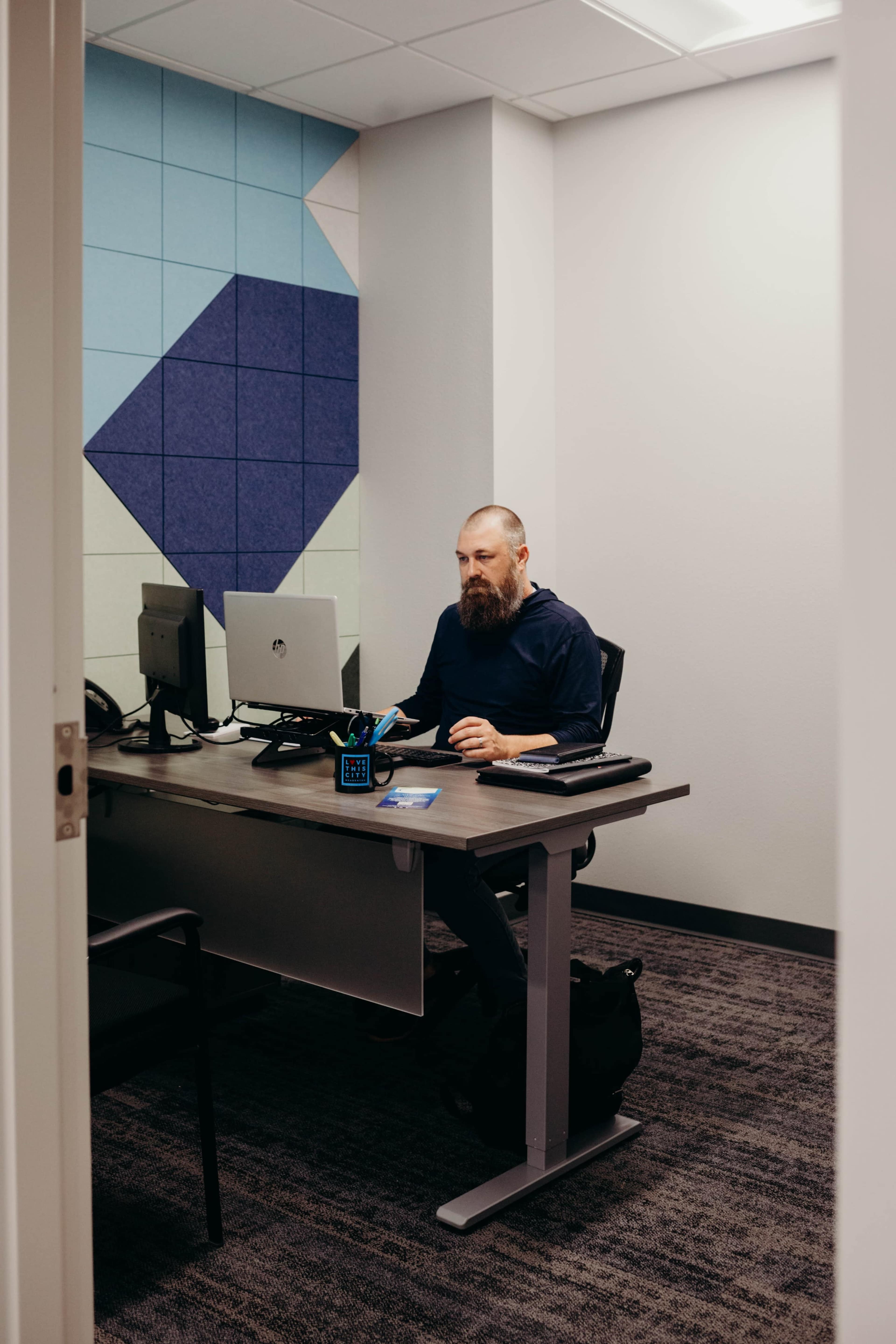 A man with a beard sits at a desk working on a computer in a modern office space with colorful wall panels.