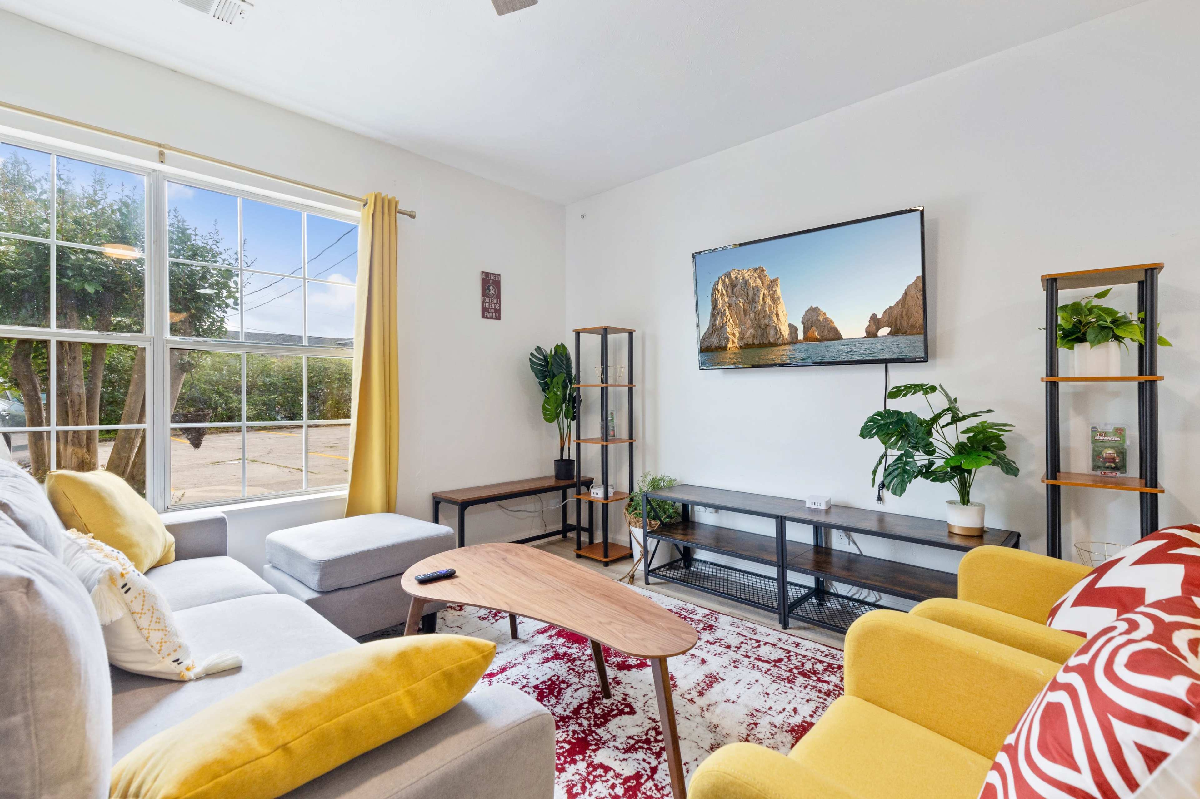 A cozy living room features a large window with yellow curtains, a sectional sofa, a wooden coffee table, a TV mounted on the wall displaying a scenic image, and decorative plants.