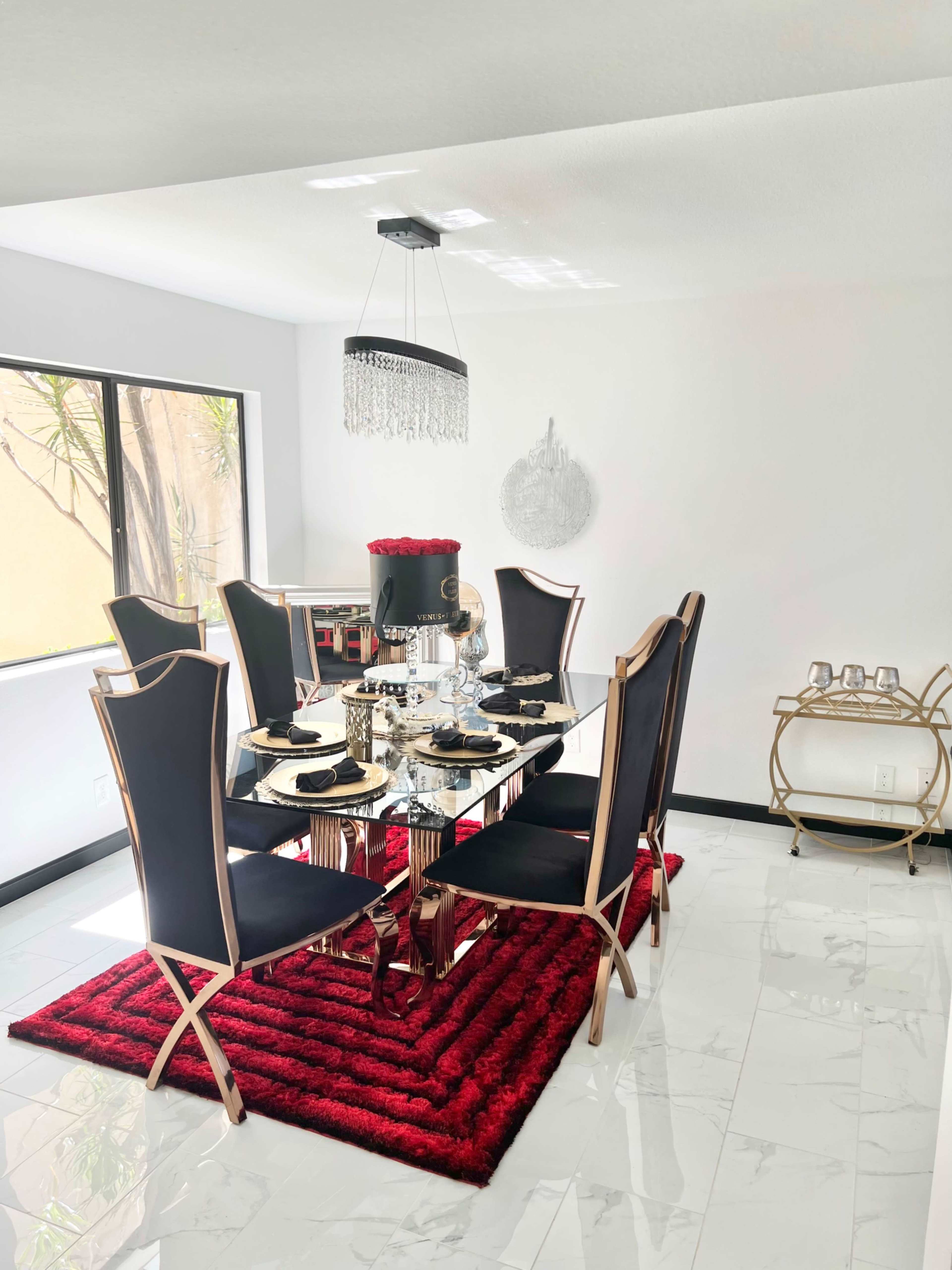 A modern dining room features a glass table surrounded by black and gold chairs, set with place settings, a red area rug, and a chandelier above.