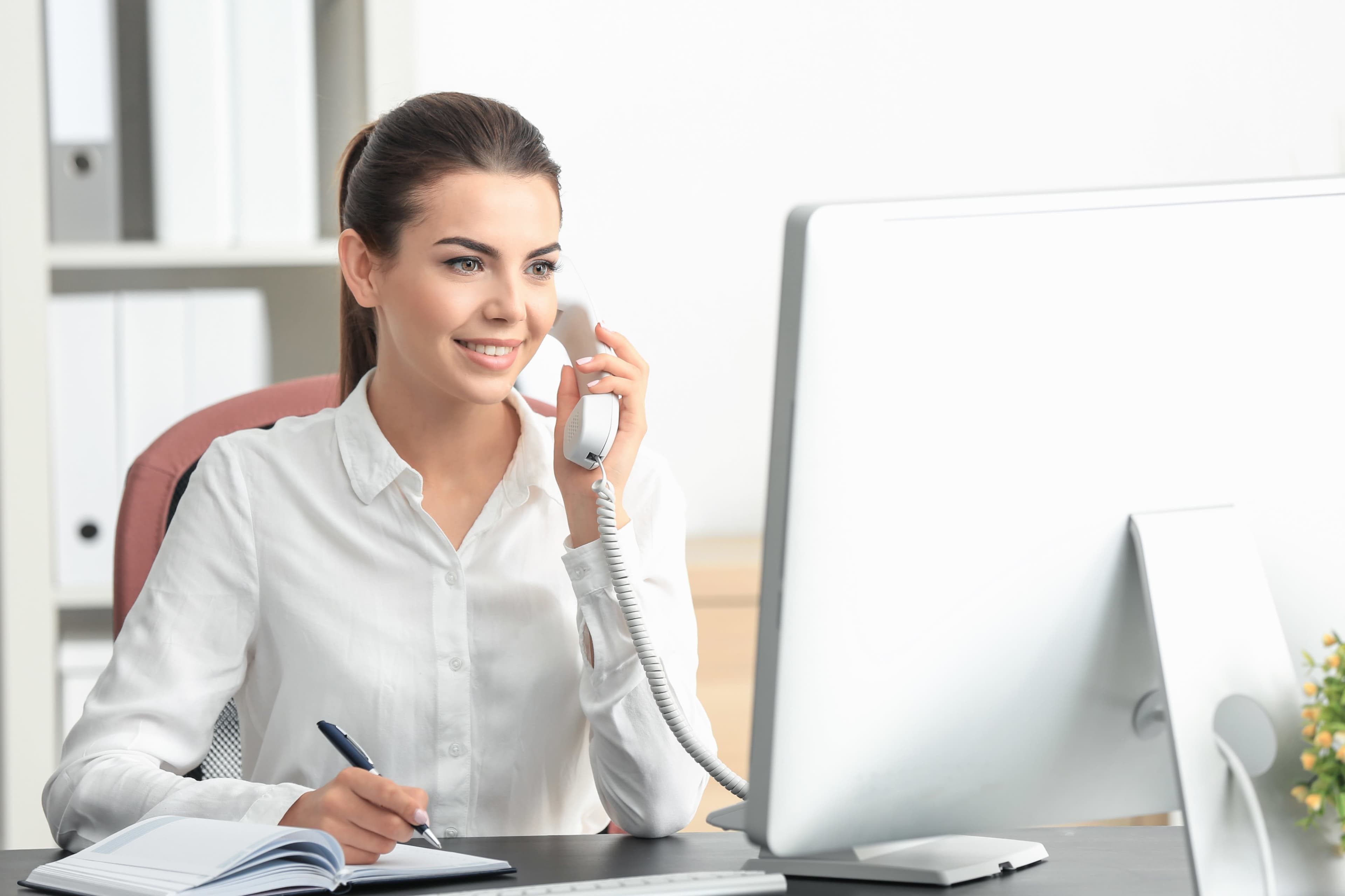 A woman is sitting at a desk, smiling while talking on a telephone and taking notes in a notebook.